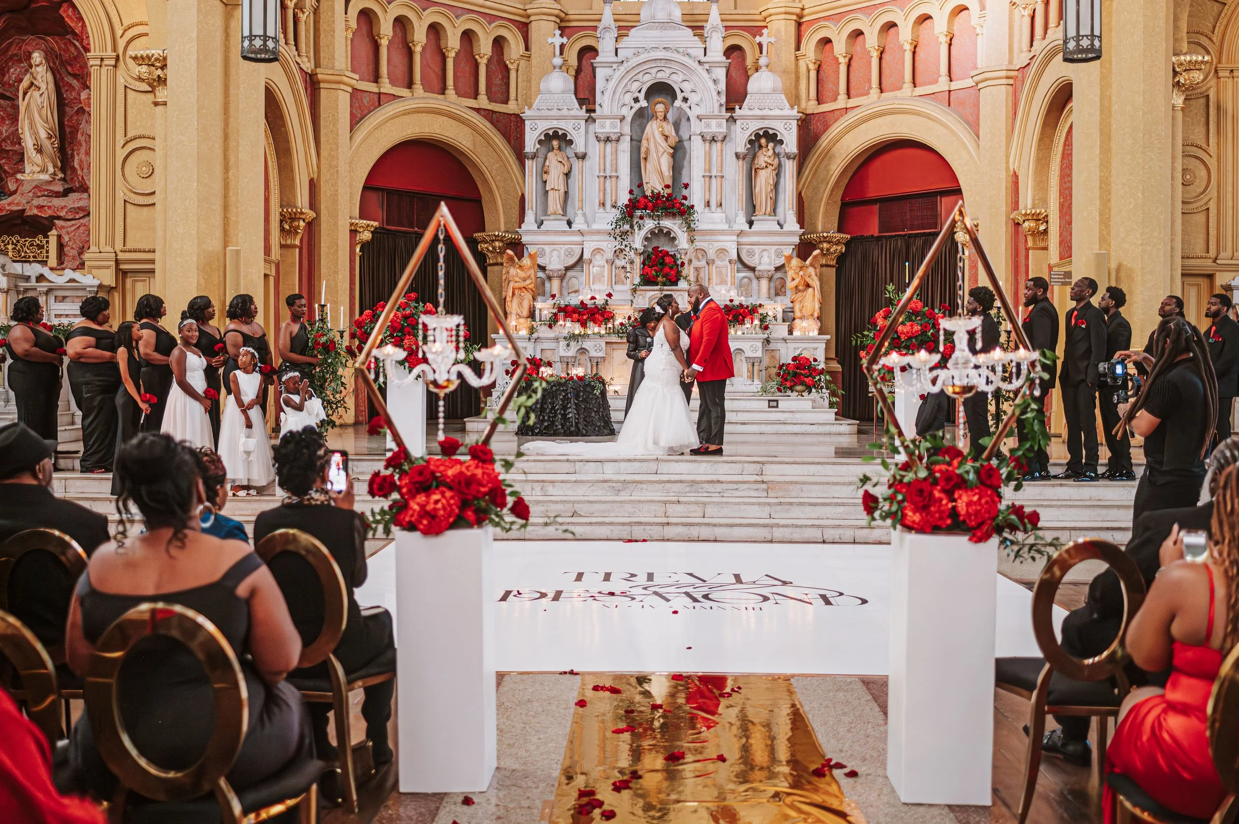 A wedding ceremony taking place inside a church with a large ornate altar decorated with statues, flowers, and red accents. The bride and groom are in the center, facing each other, with guests seated on either side and women and children dressed in 