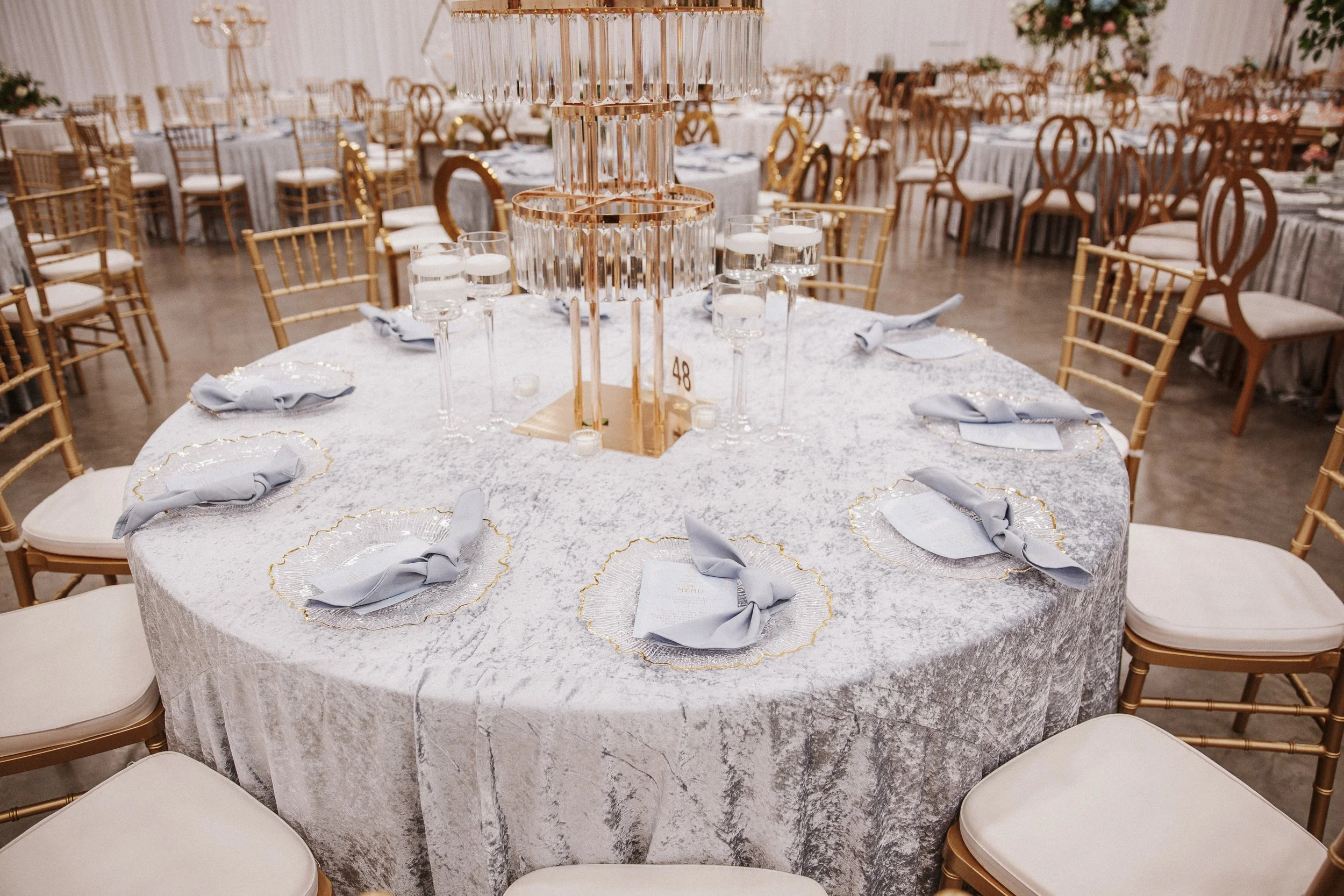 Elegant banquet hall with round table covered in a silver tablecloth, set with gray napkins, menu cards, and glassware. Gold chairs surround the table, and a large, crystal chandelier hangs above.
