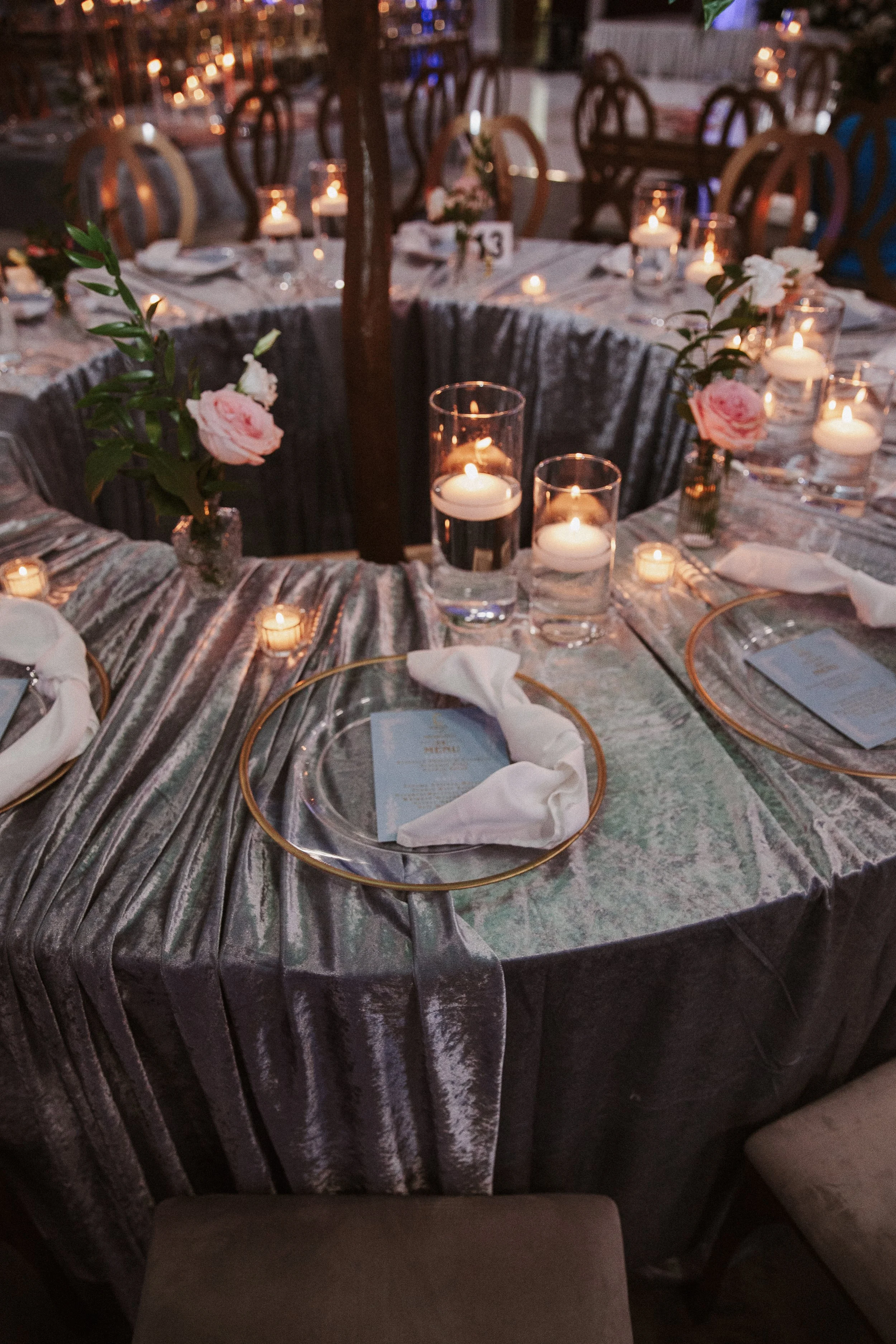 A round dining table covered with a silver tablecloth, set with candles in glass holders, pink roses, and place settings. There are chairs around the table, and the scene is lit with candlelight, suggesting an evening event or celebration.