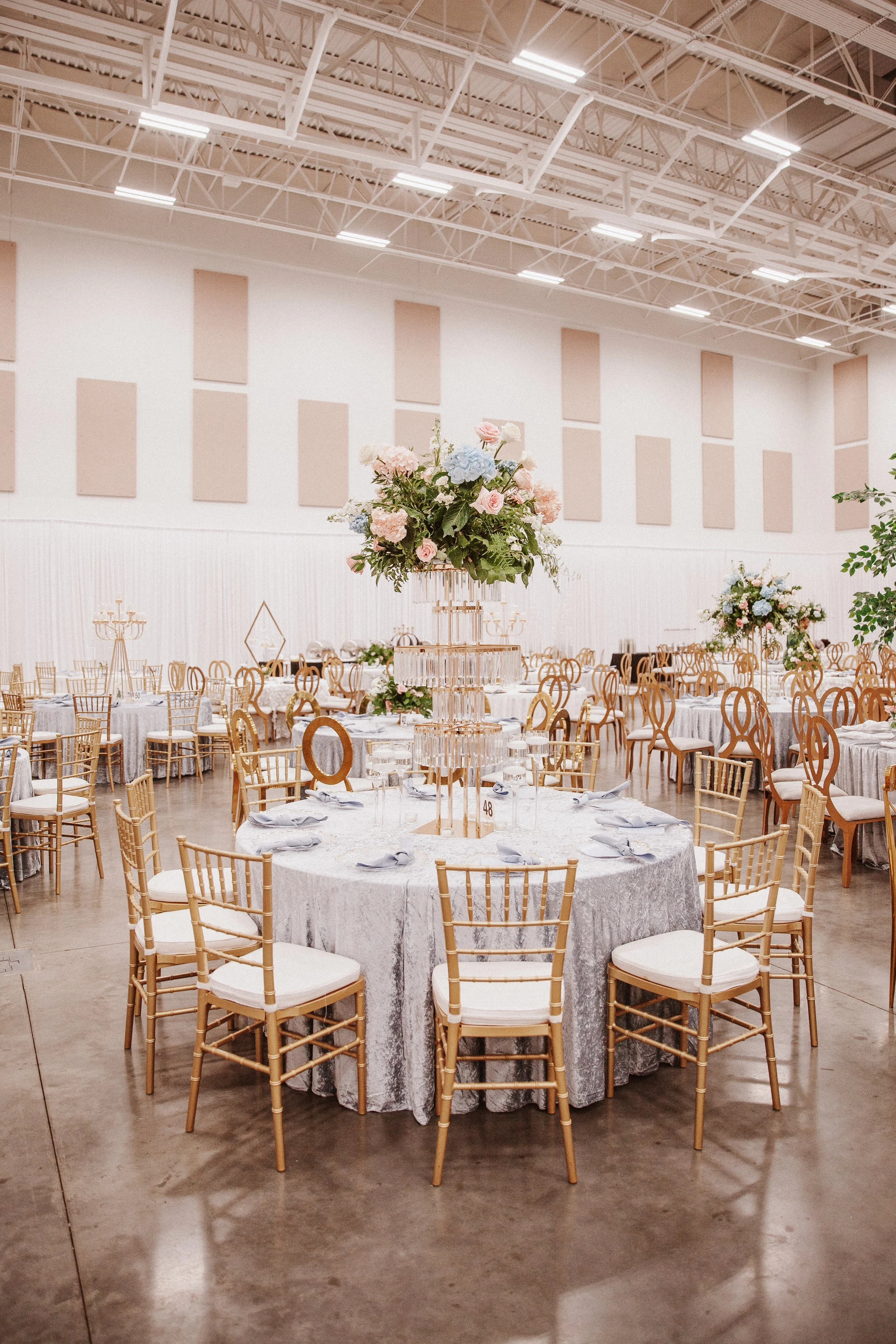 A banquet hall decorated for an event with round tables covered in silver tablecloths, adorned with floral centerpieces, and surrounded by gold chairs with white cushions. The hall has a high ceiling with exposed metal beams and soft pink sound-absor