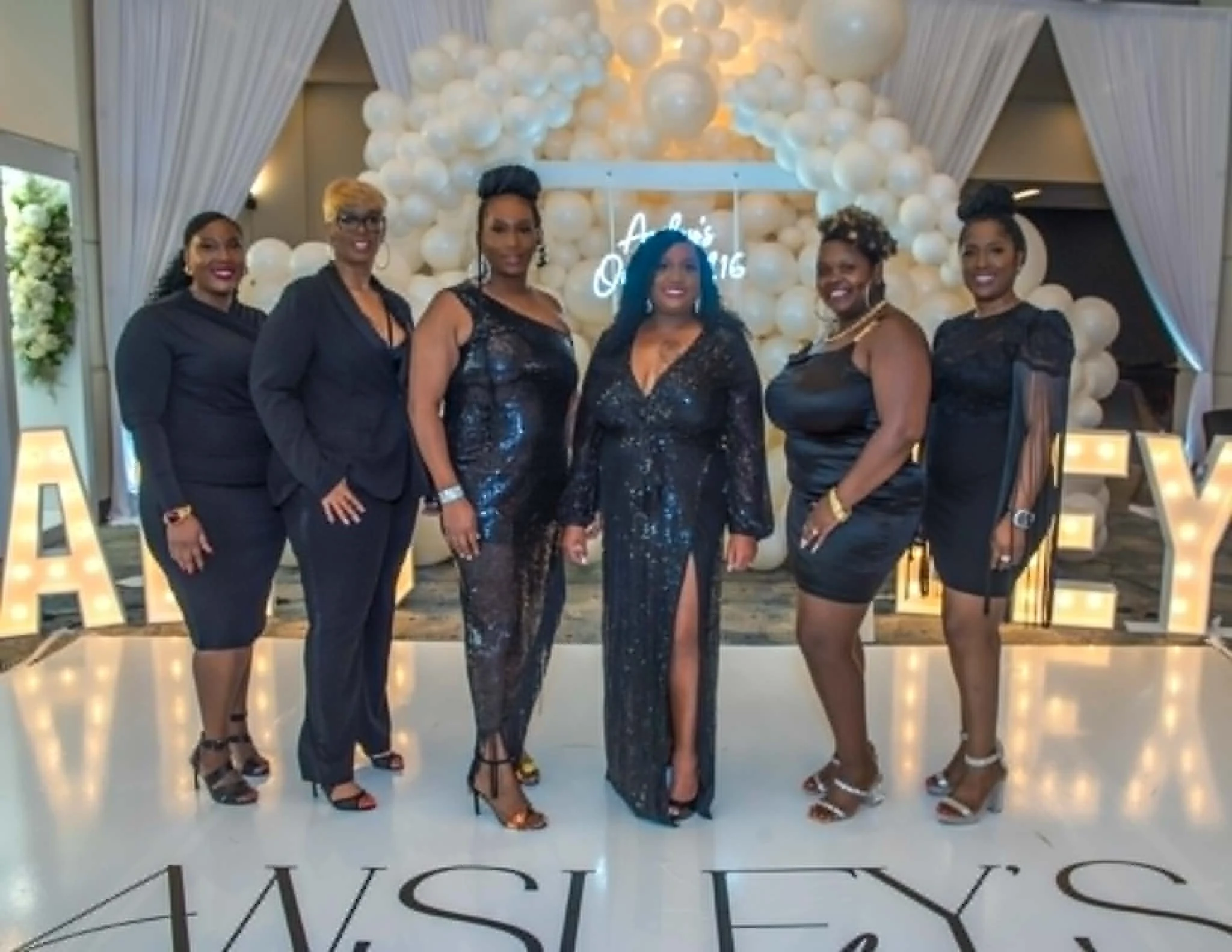 Six women dressed in black dresses and high heels pose together at an event with an elegant backdrop of white balloons, illuminated letters, and draped curtains.