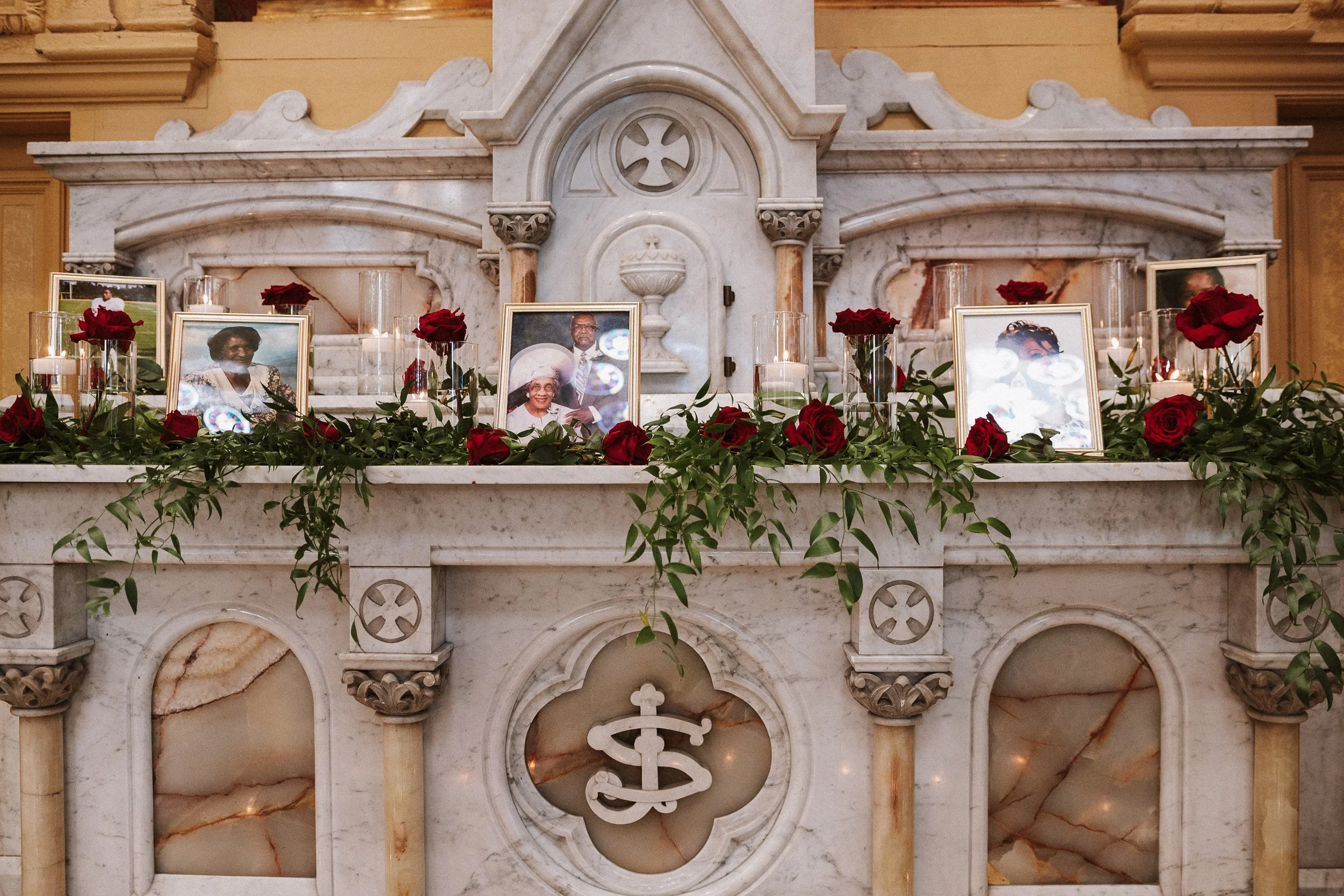 Memorial altar with framed photographs of loved ones, red roses in glass vases, and lit candles, set against a marble backdrop with religious symbols.