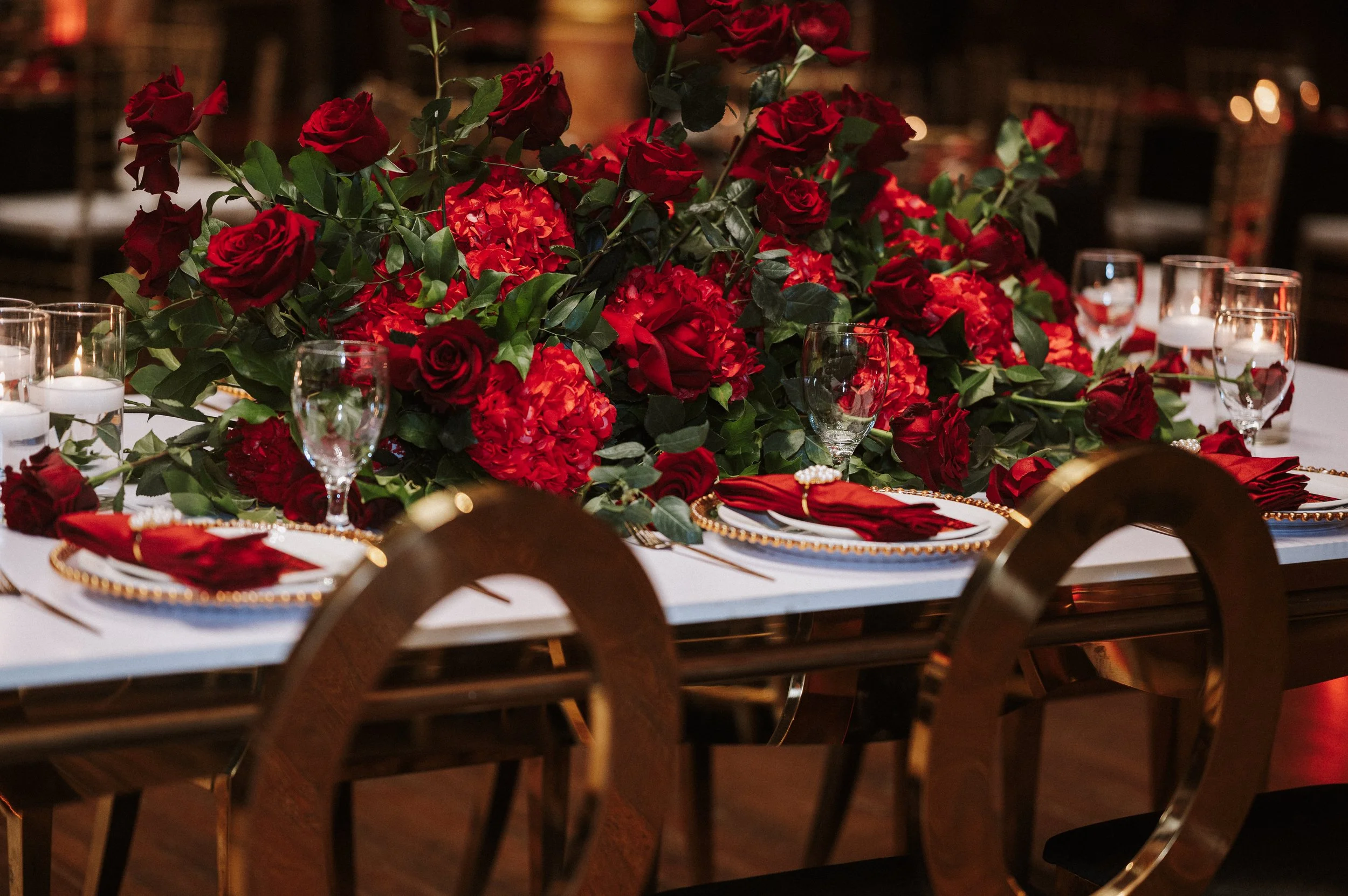 A beautifully decorated table with a large bouquet of red and orange flowers, surrounded by wine glasses, candles, and elegant table settings, in a warm, dimly lit environment.
