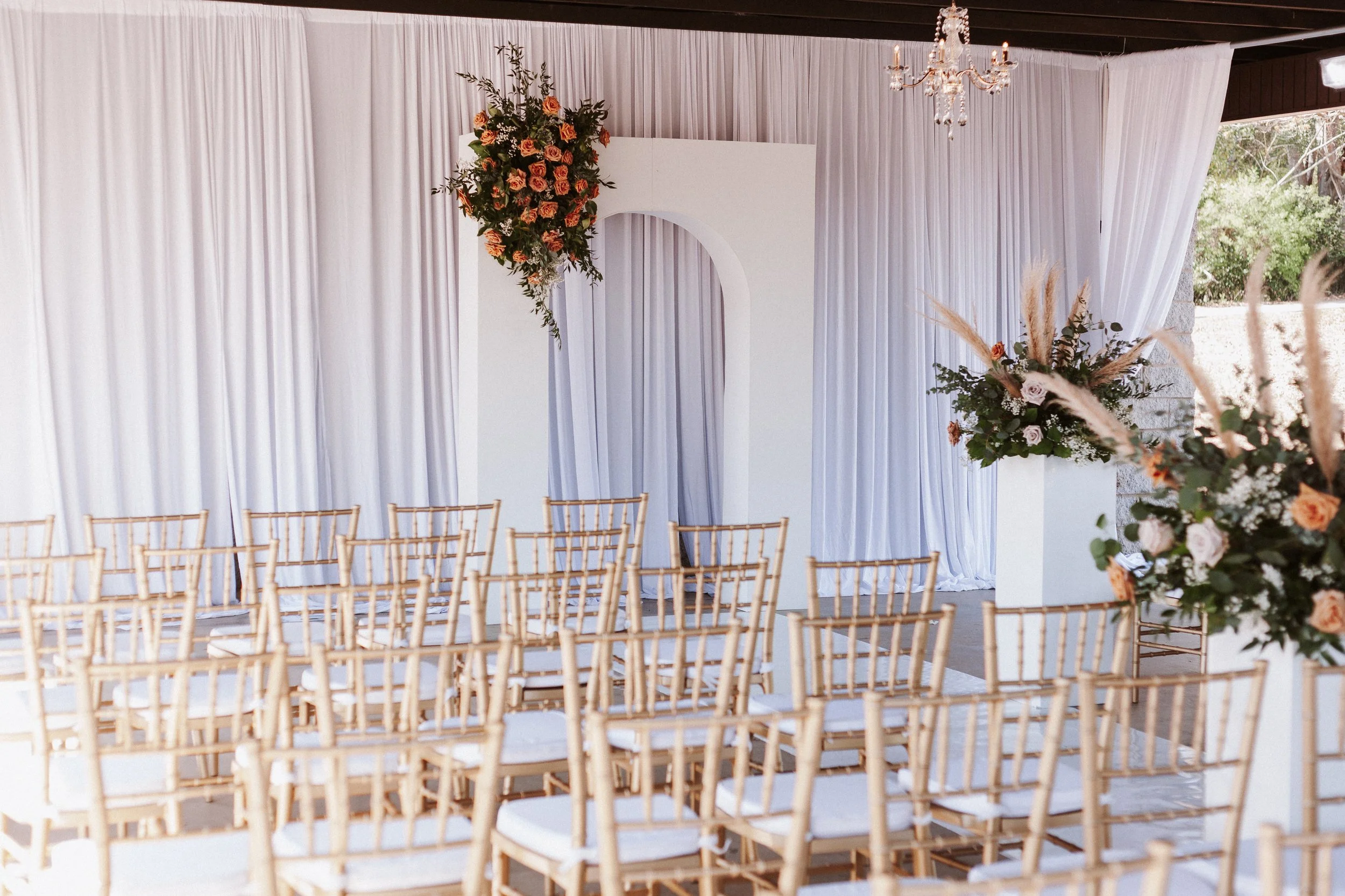 Wedding ceremony setup with rows of gold chairs facing a white backdrop with floral arrangements, a white arch, and a chandelier overhead.