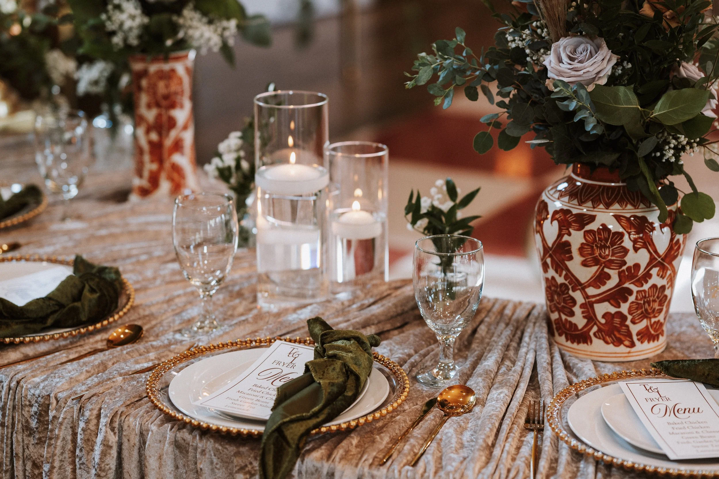 A decorated dining table with a floral centerpiece in a patterned vase, several candles in glass holders, place settings with gold beaded chargers, green napkins, and menus, on a textured tablecloth.
