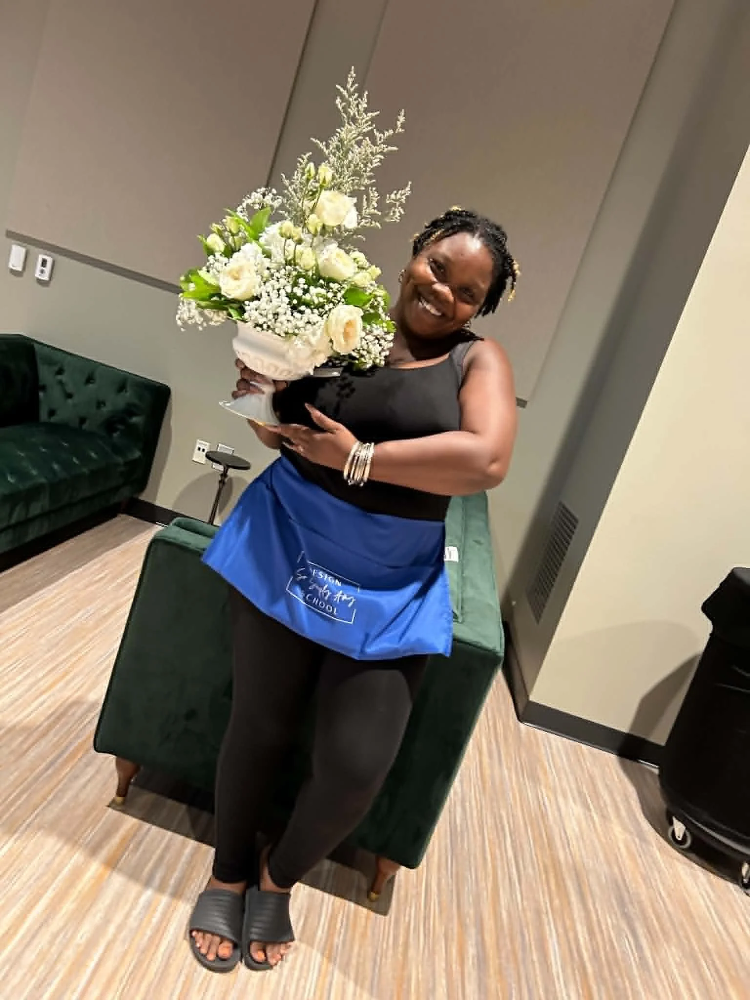A woman smiling and holding a large bouquet of white flowers while seated on a green velvet chair in a room with tan carpet, gray walls, and various furniture and decor.
