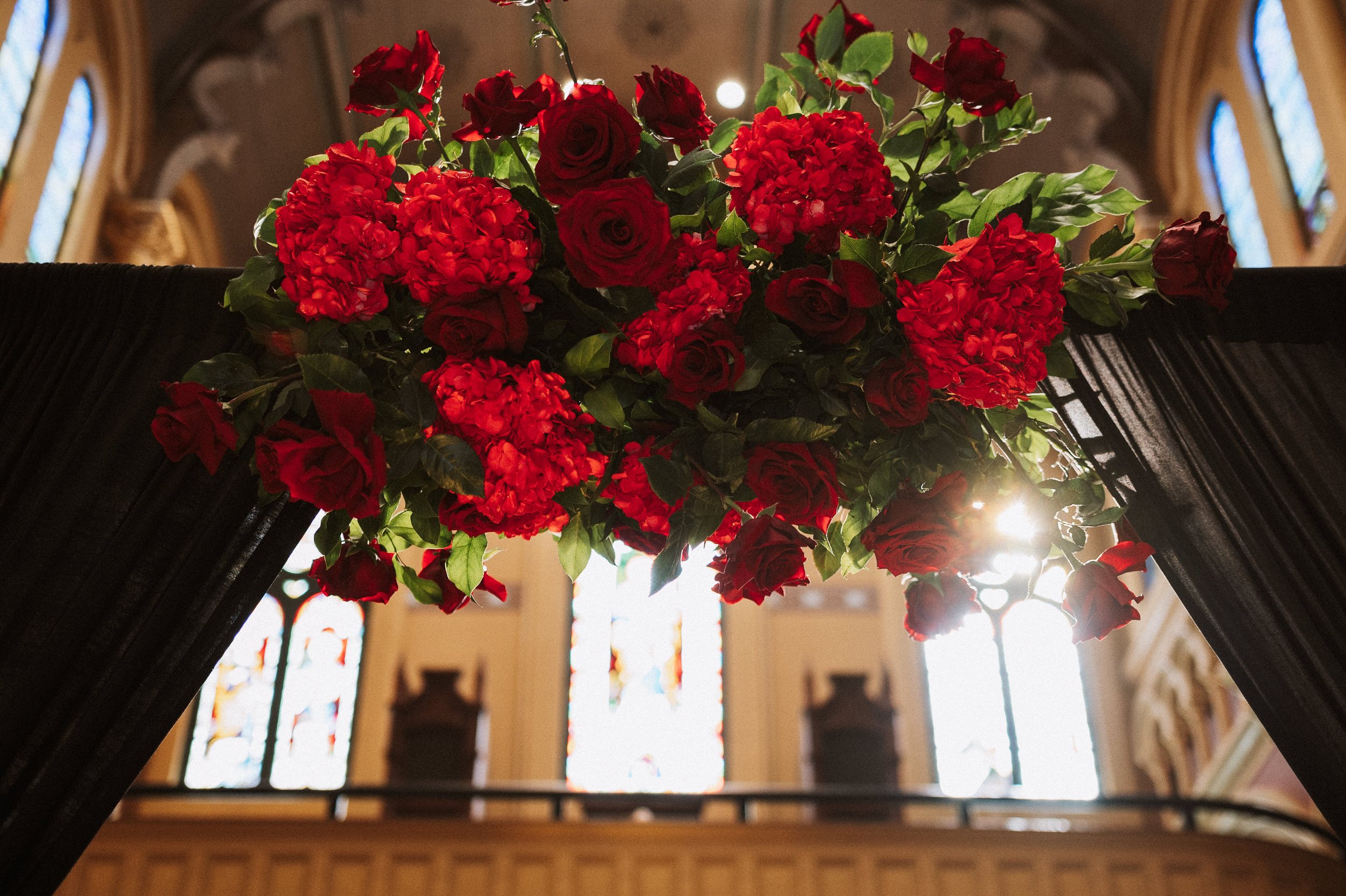 A large bouquet of red roses, hydrangeas, and greenery atop a black piano inside a church with stained glass windows in the background.