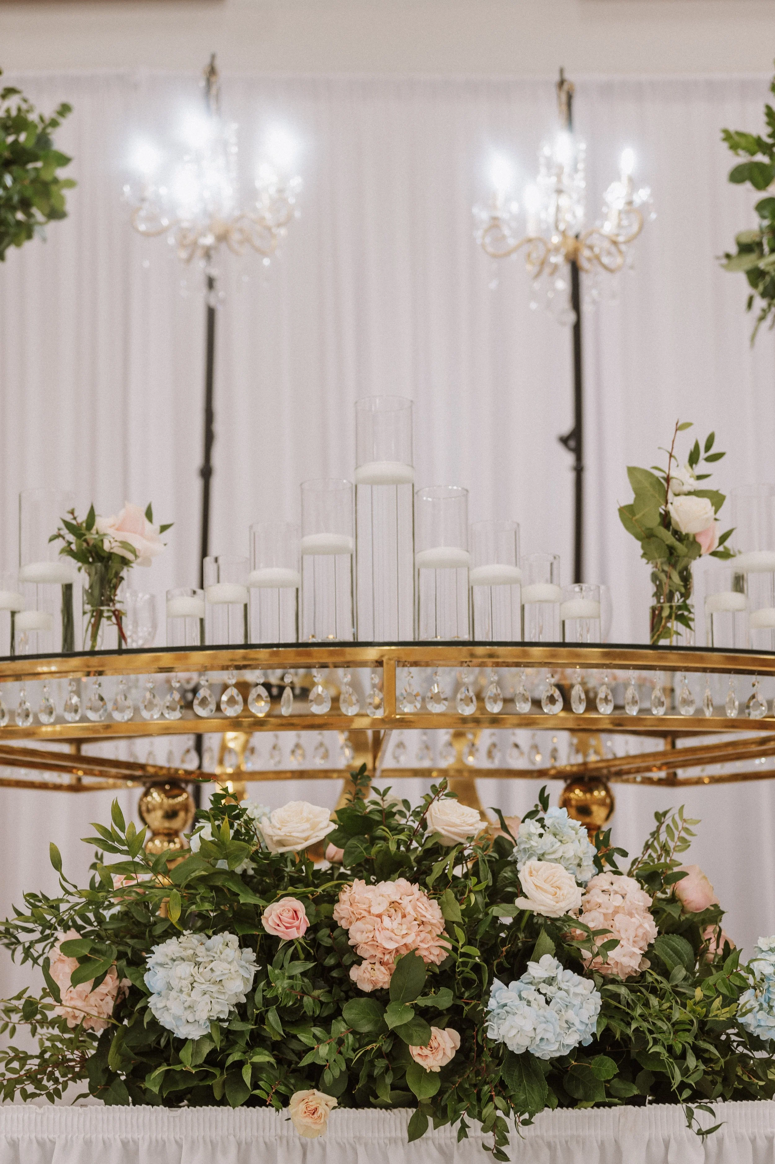 Elegant floral arrangement with pink, white, and blue hydrangeas, roses, and greenery on a gold and crystal stand at a wedding or event.