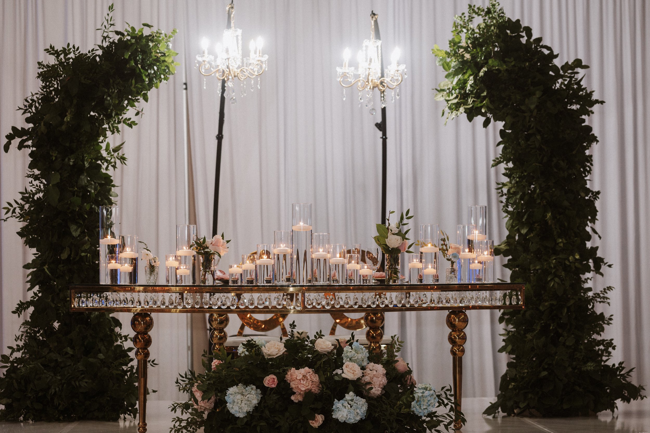 A decorated table with lit candles, surrounded by floral arrangements and greenery, set against a white curtain backdrop with chandeliers overhead.