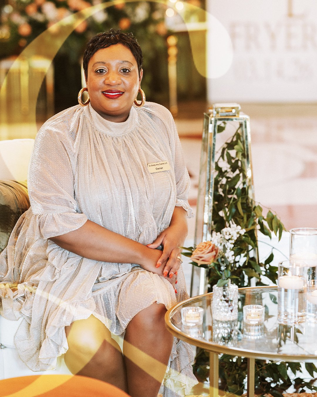 A woman sitting on a white chair, wearing a beige dress with ruffled sleeves and gold hoop earrings, smiling at the camera. She is indoors next to a glass table with candles and a small floral arrangement, with a name tag reading 'Owner' on her dress.
