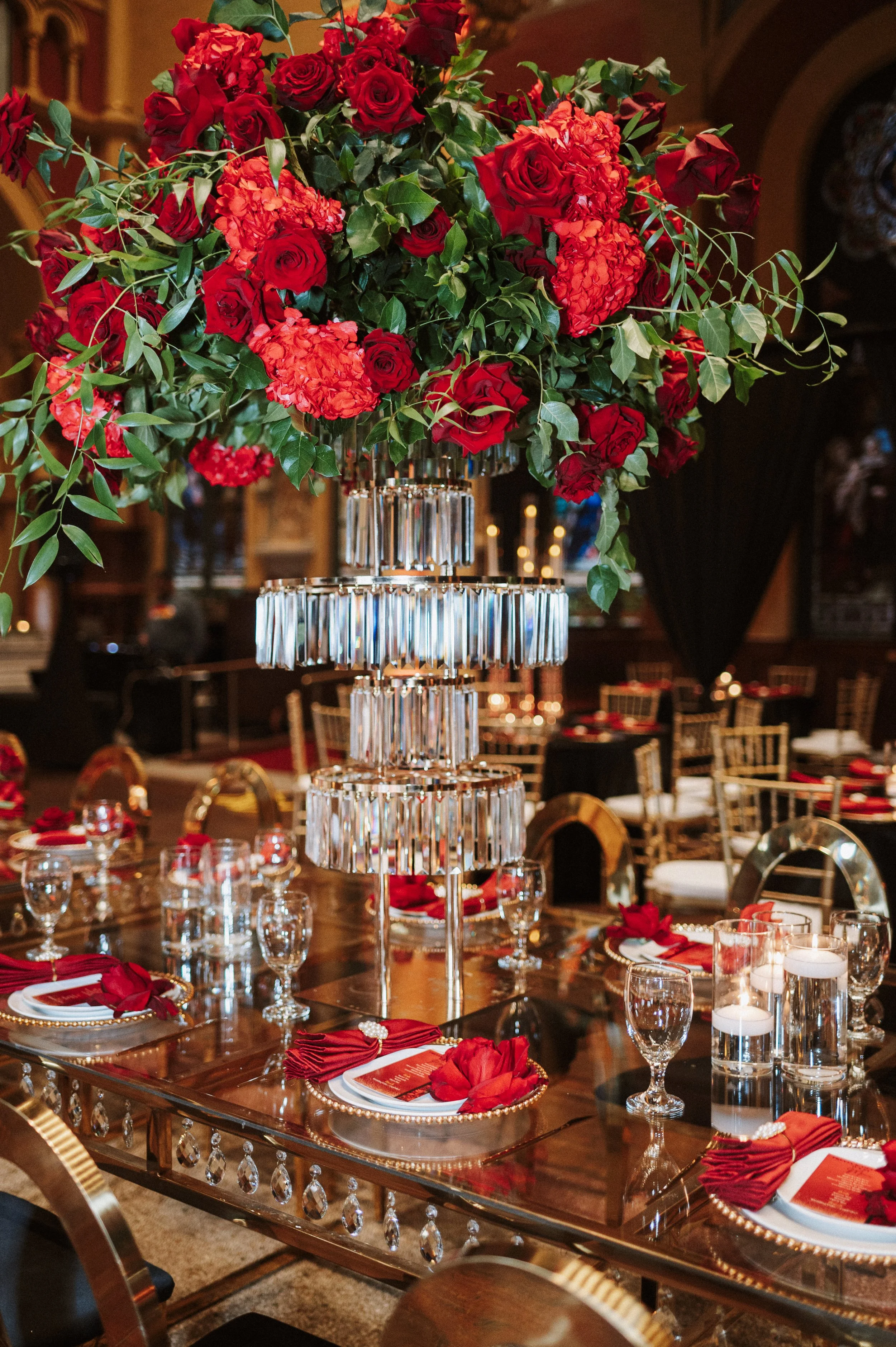 An elegant table setting with a large floral centerpiece consisting of red roses and red hydrangeas, surrounded by candles in glass holders and red napkins on plates, in a decorated dining room with chandeliers and golden chairs.