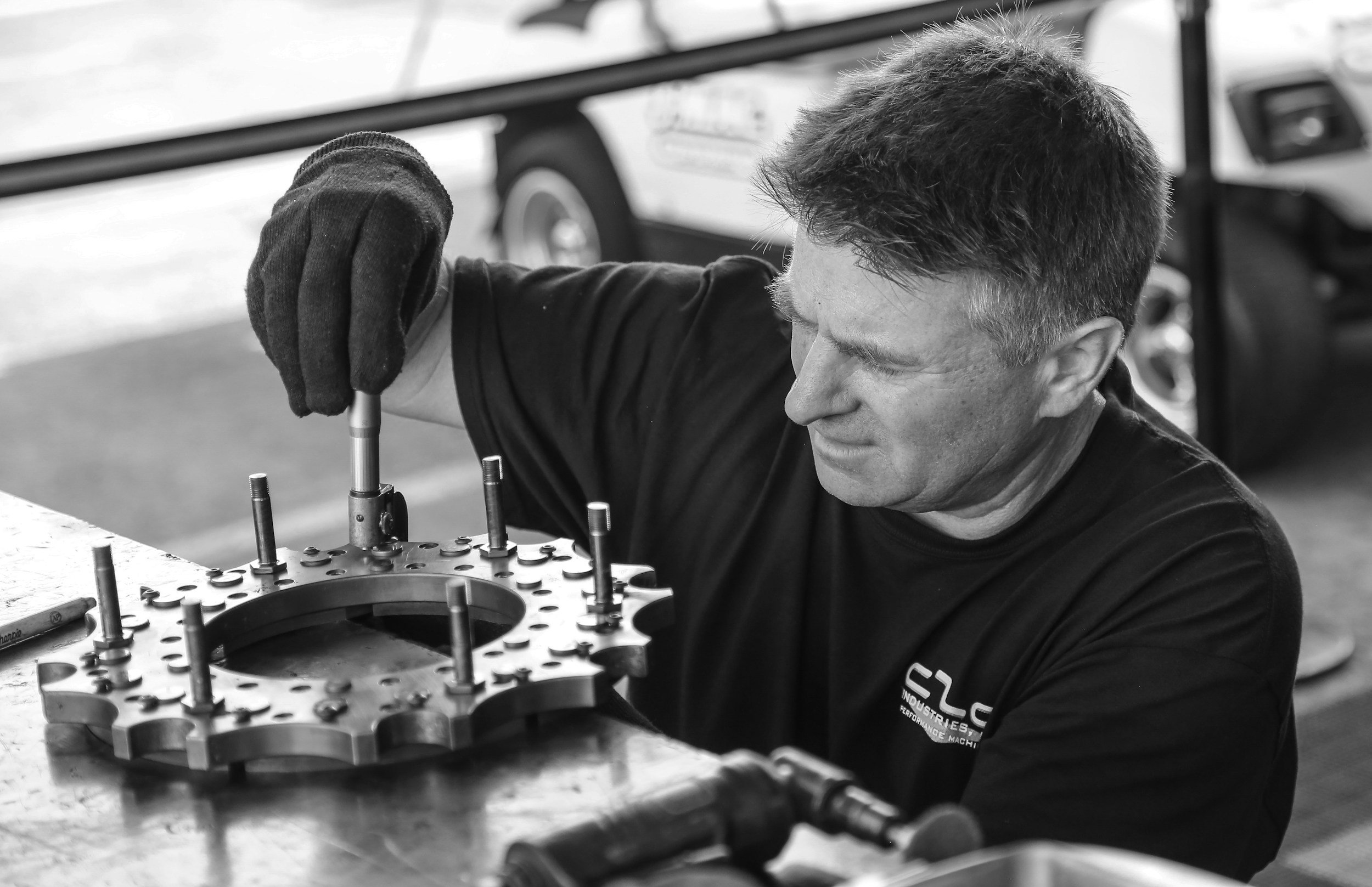 A man wearing gloves and a dark shirt working on an engine component with a circular metal part and vertical pins, in a workshop setting.