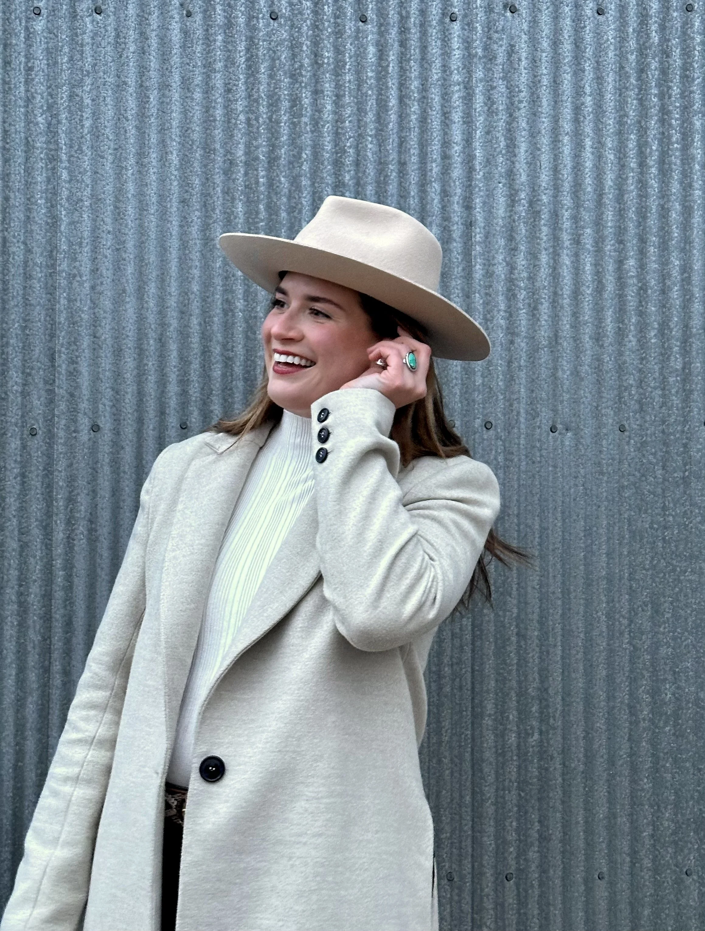 A young woman wearing a beige blazer and a white turtleneck, smiling and holding her right hand to her ear, standing in front of a corrugated metal wall, wearing a wide-brimmed beige hat and a turquoise ring.