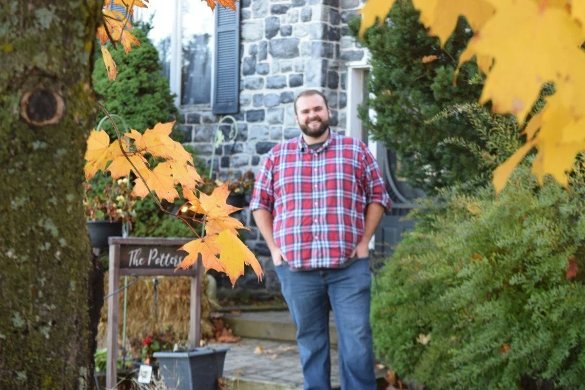 A man with a beard and short hair, wearing a red plaid shirt and jeans, standing outdoors in front of a stone house surrounded by trees with yellow and orange fall leaves.
