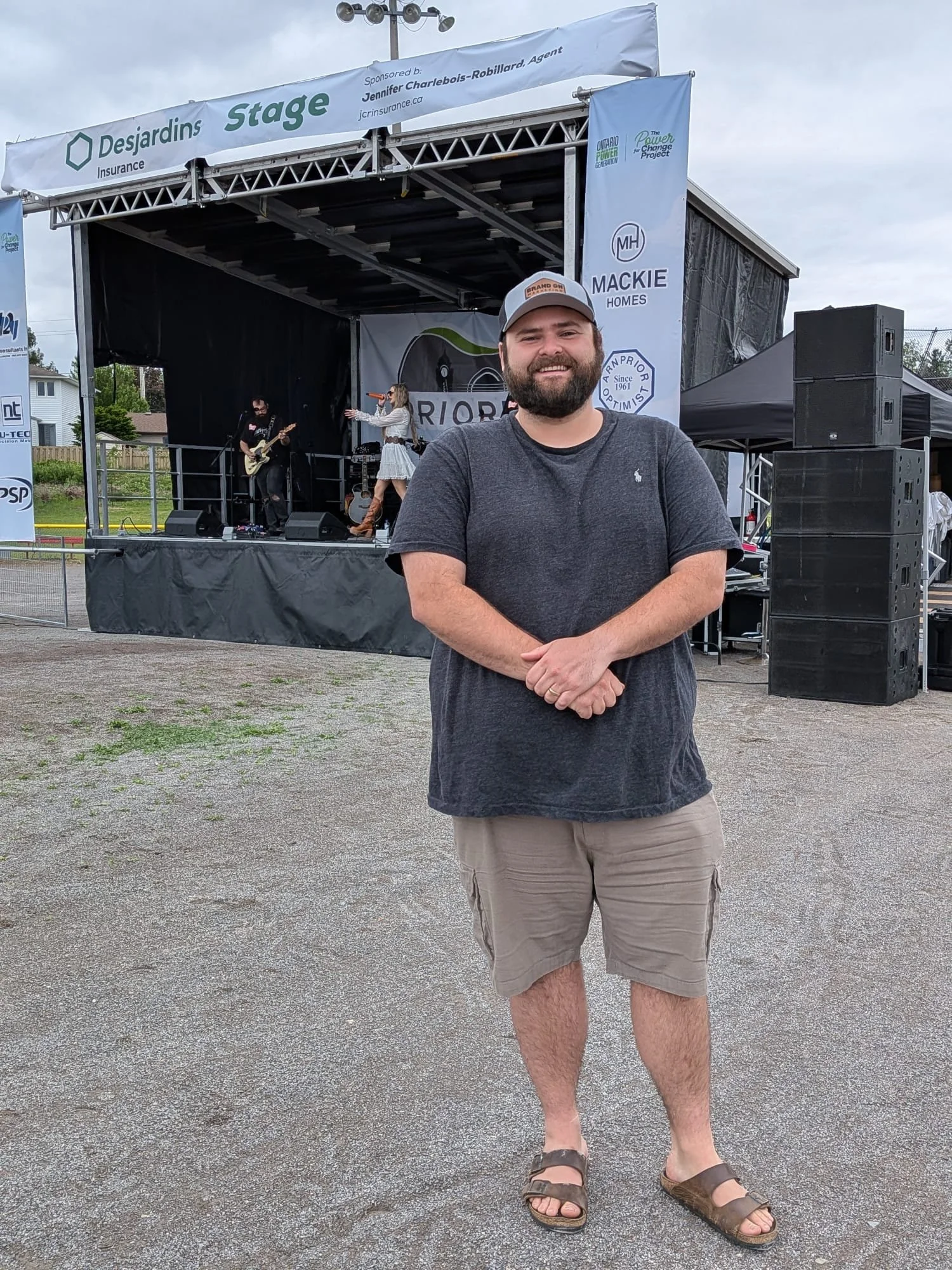 A man standing in front of an outdoor stage with a band performing. He is smiling, wearing a gray t-shirt, beige shorts, sandals, and a cap. The stage has banners and speakers, with cloudy skies overhead.
