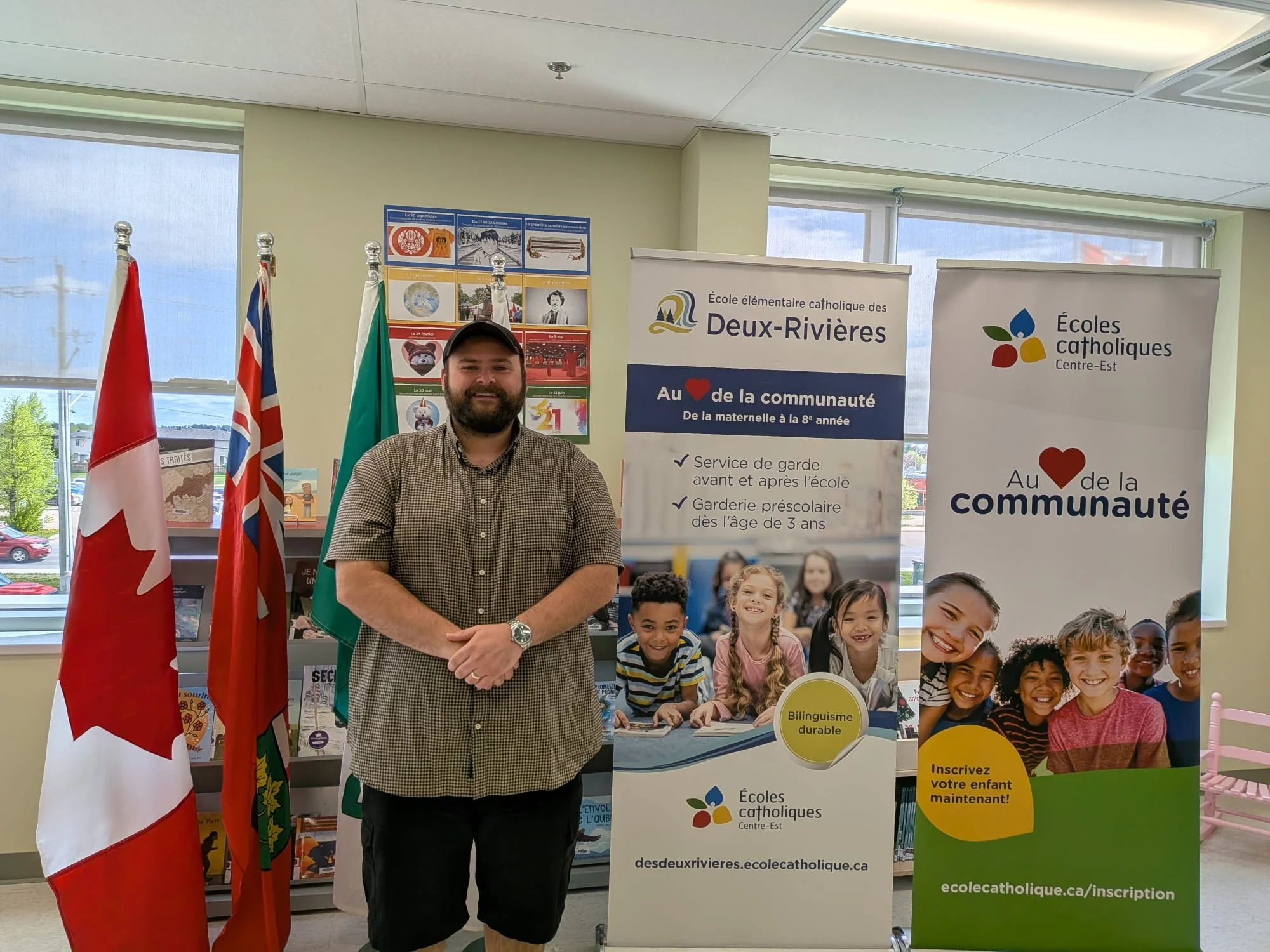 A man with a beard, wearing a checkered shirt and black shorts, standing indoors in front of promotional banners and flags. The banners promote 'Écoles catholiques' and the community of Deux-Rivières, with images of children and information about bilingual education and registration. The background includes windows, colorful posters, and greenery outside.