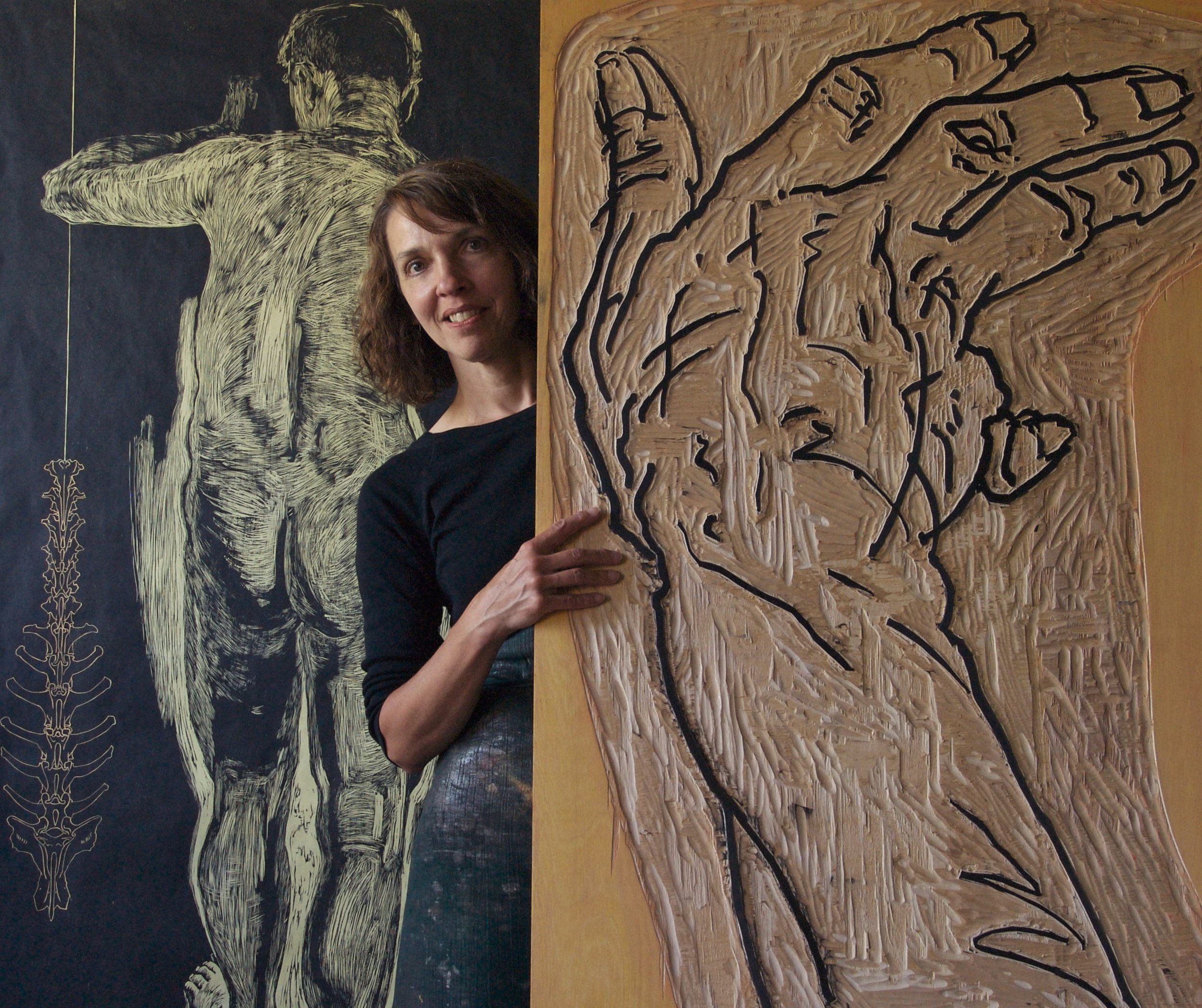 A woman standing in front of two artworks. One on the left is a large black background with a chalk-like sketch of a human figure with outstretched arms. The artwork on the right is a textured abstract piece with black lines on a beige background, resembling an anatomical or organic form.