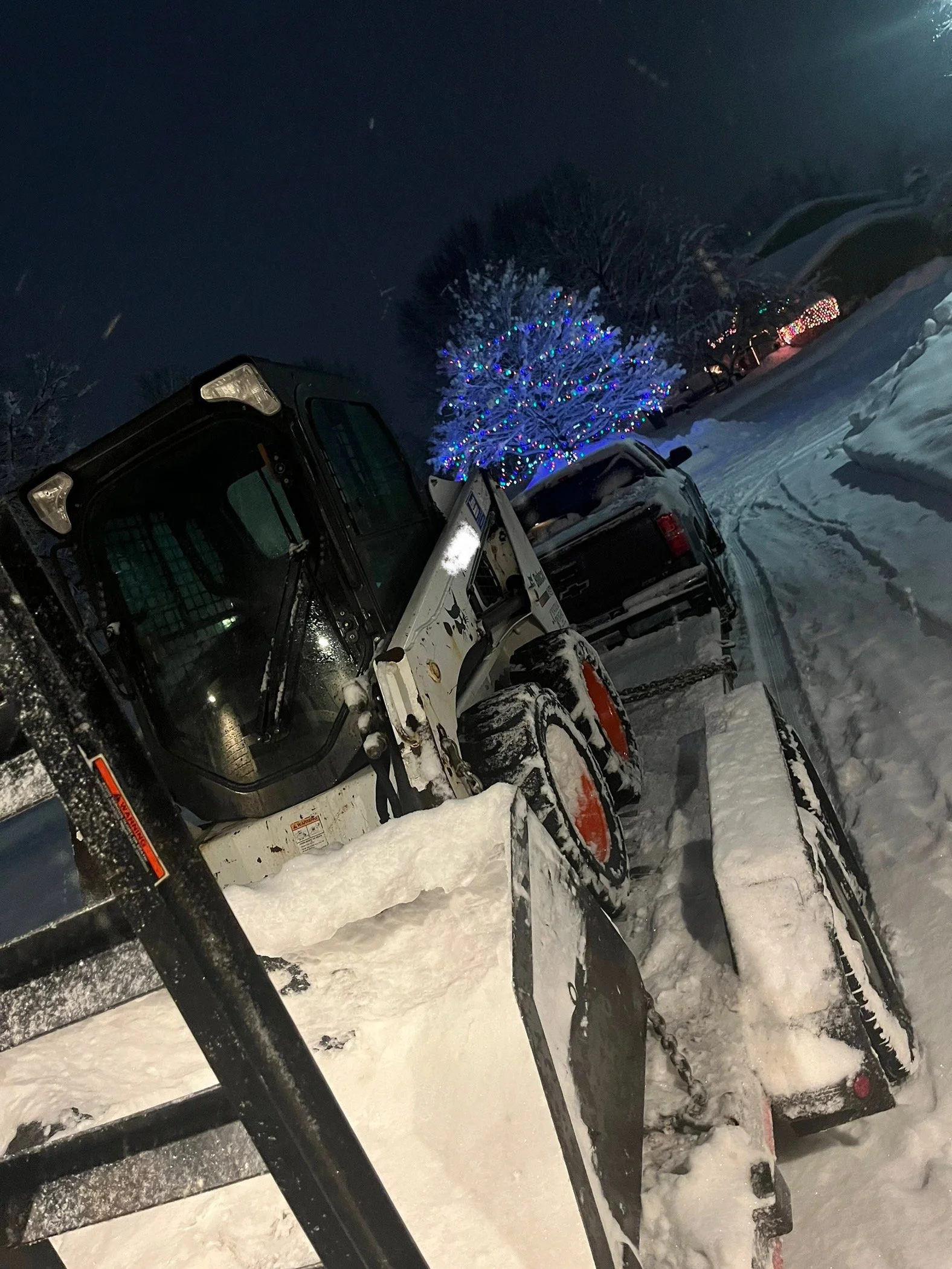 Snow-covered driveway with a skid-steer loader clearing snow, a snow-covered truck parked nearby, and a tree decorated with colorful Christmas lights in the background at night.