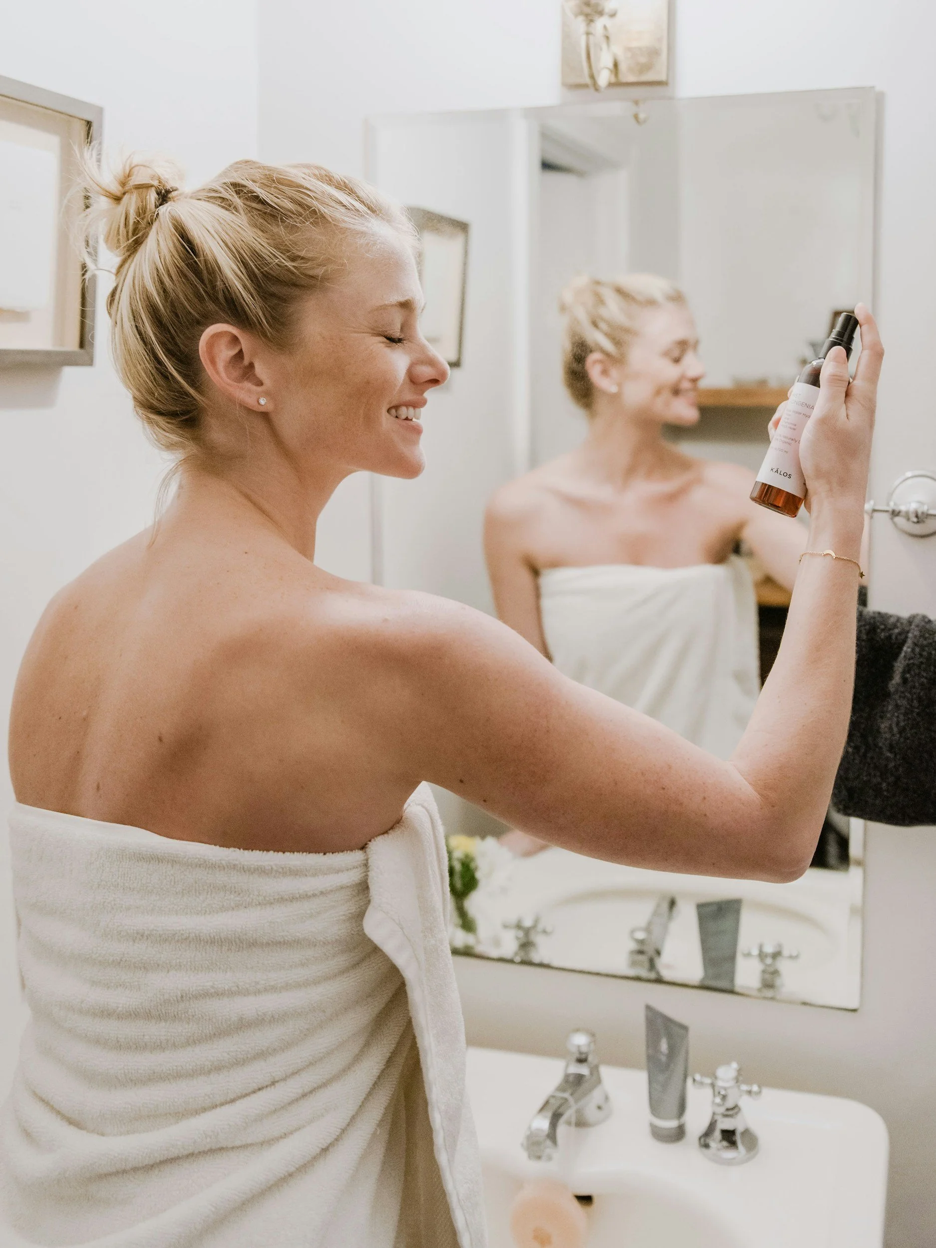 A woman smiling with her eyes closed, wrapped in a towel, holding a spray bottle in a bathroom mirror performing part of her skincare routine.