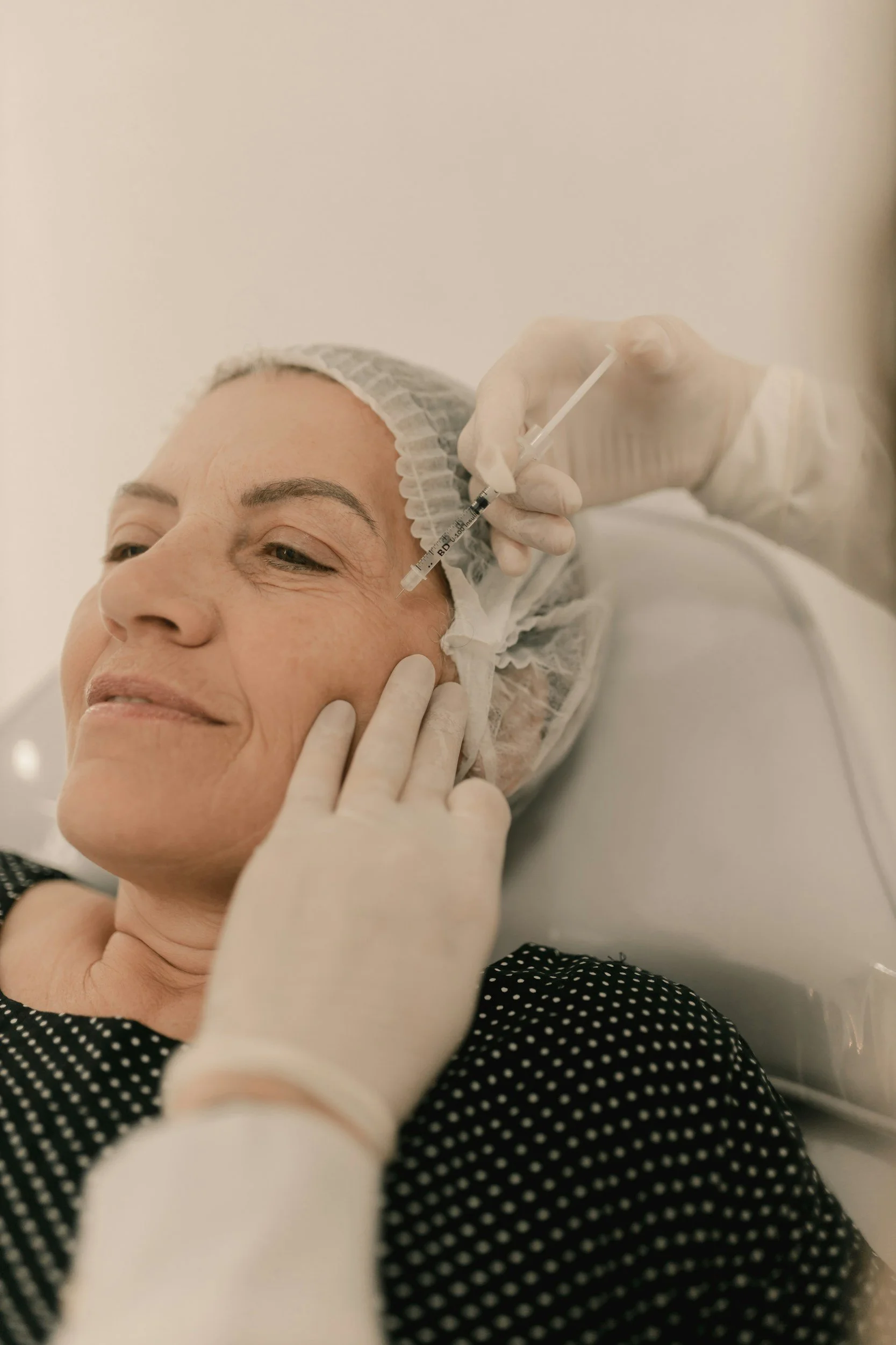 Close-up of a woman lying on a medical treatment bed, receiving a facial injection from a healthcare professional wearing gloves, in a clinical setting.