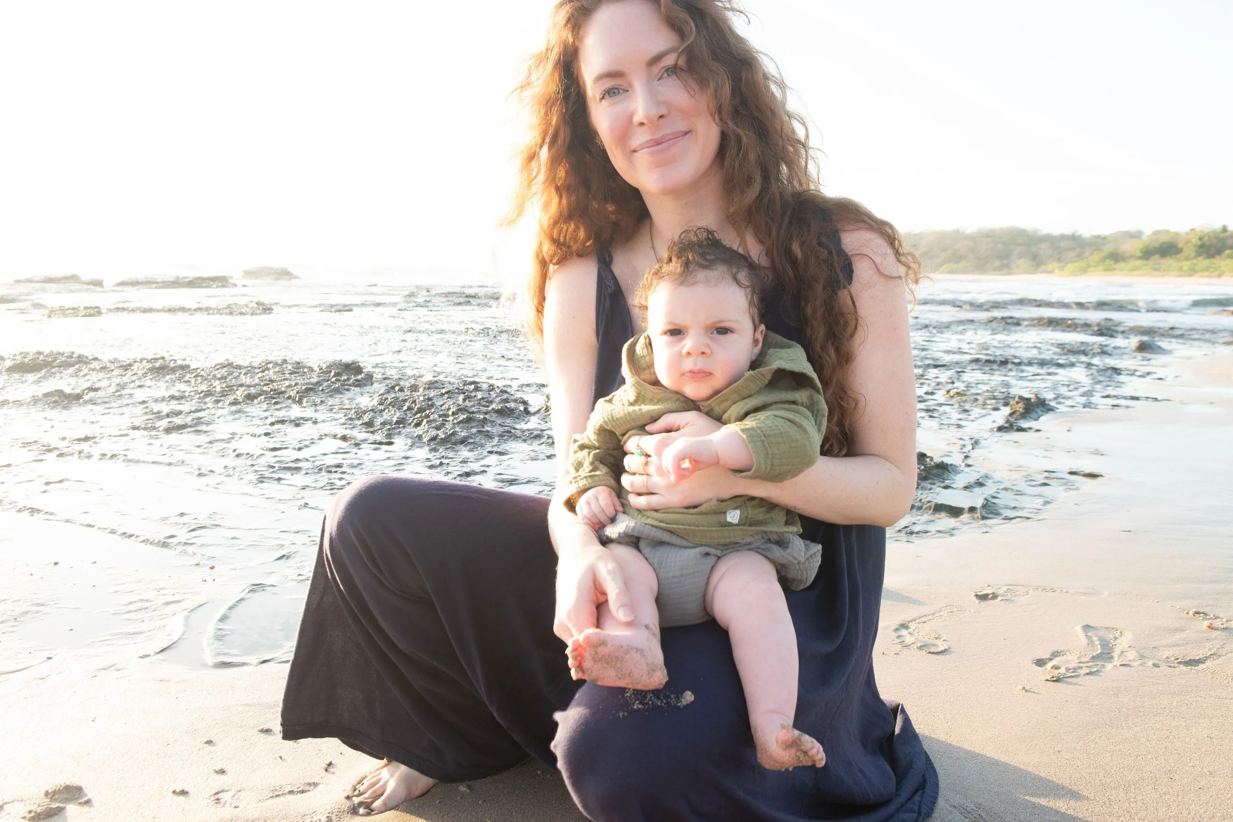 A woman with long curly brown hair holding a young child with short curly hair on a beach at sunset, with ocean waves and a distant shoreline in the background.