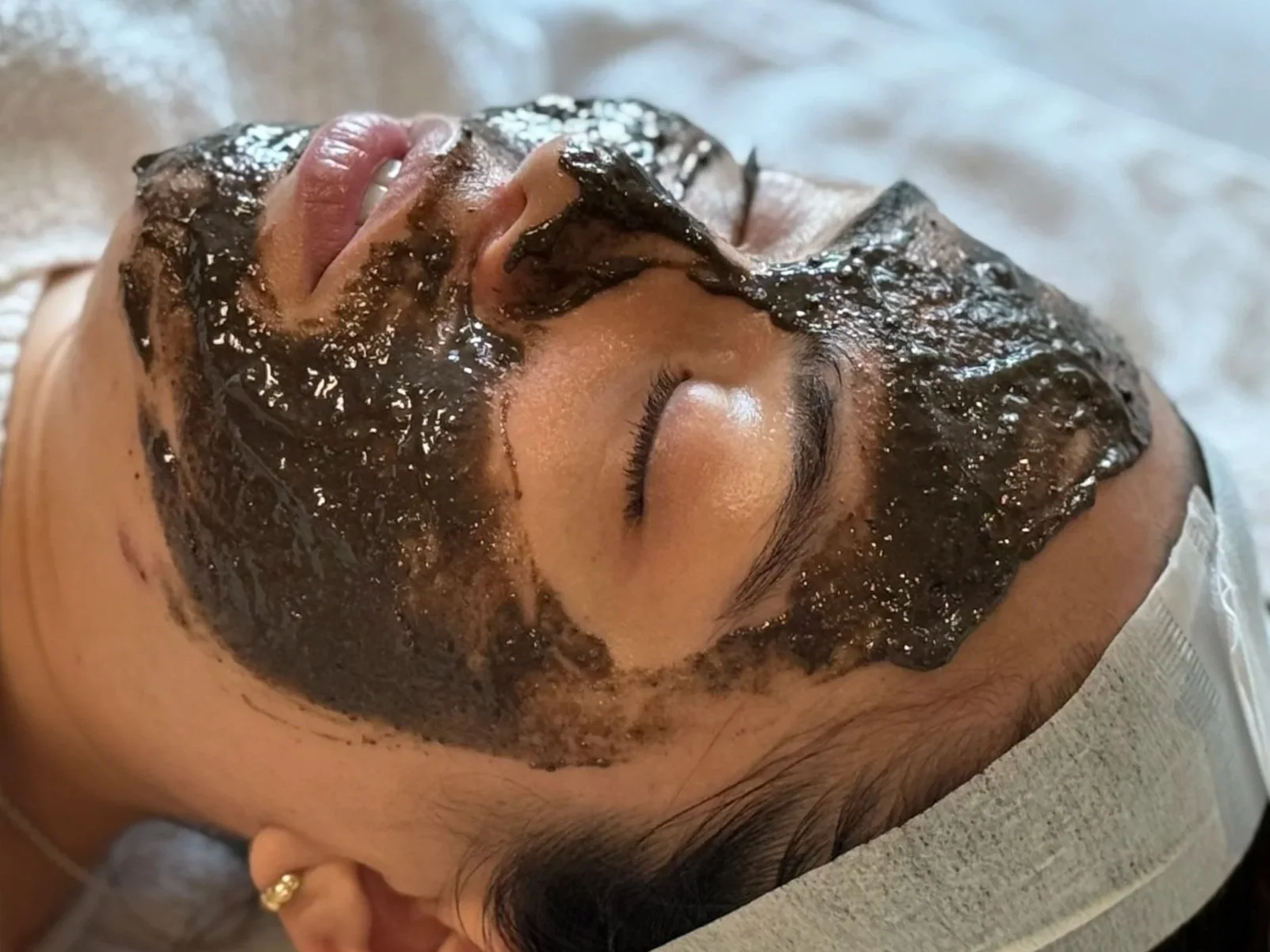 A woman lying down with her eyes closed, receiving a facial mask treatment. The mask is dark-colored charcoal and applied evenly on her face.