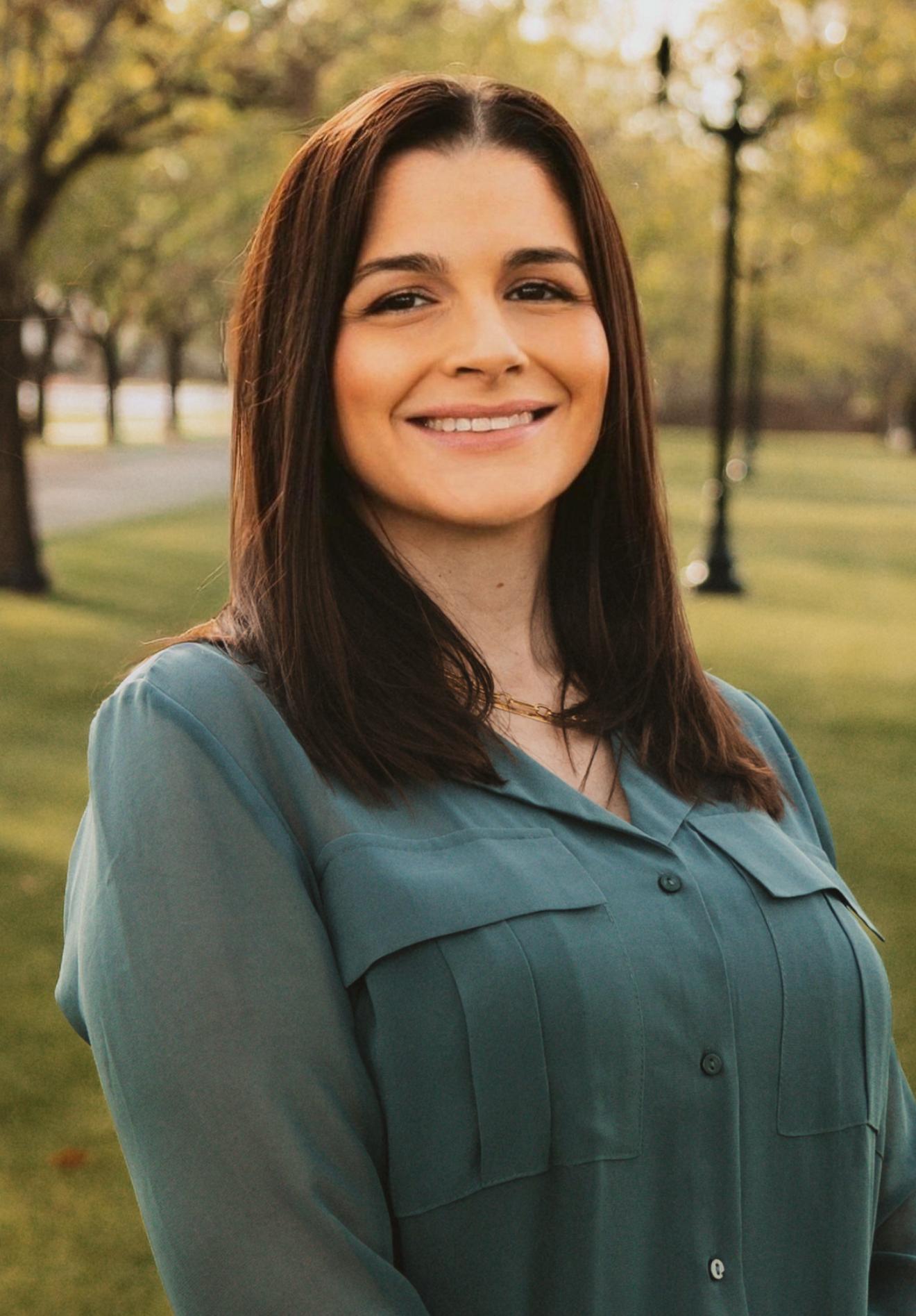 A woman with long dark hair is smiling outdoors with trees and a park-like setting in the background.