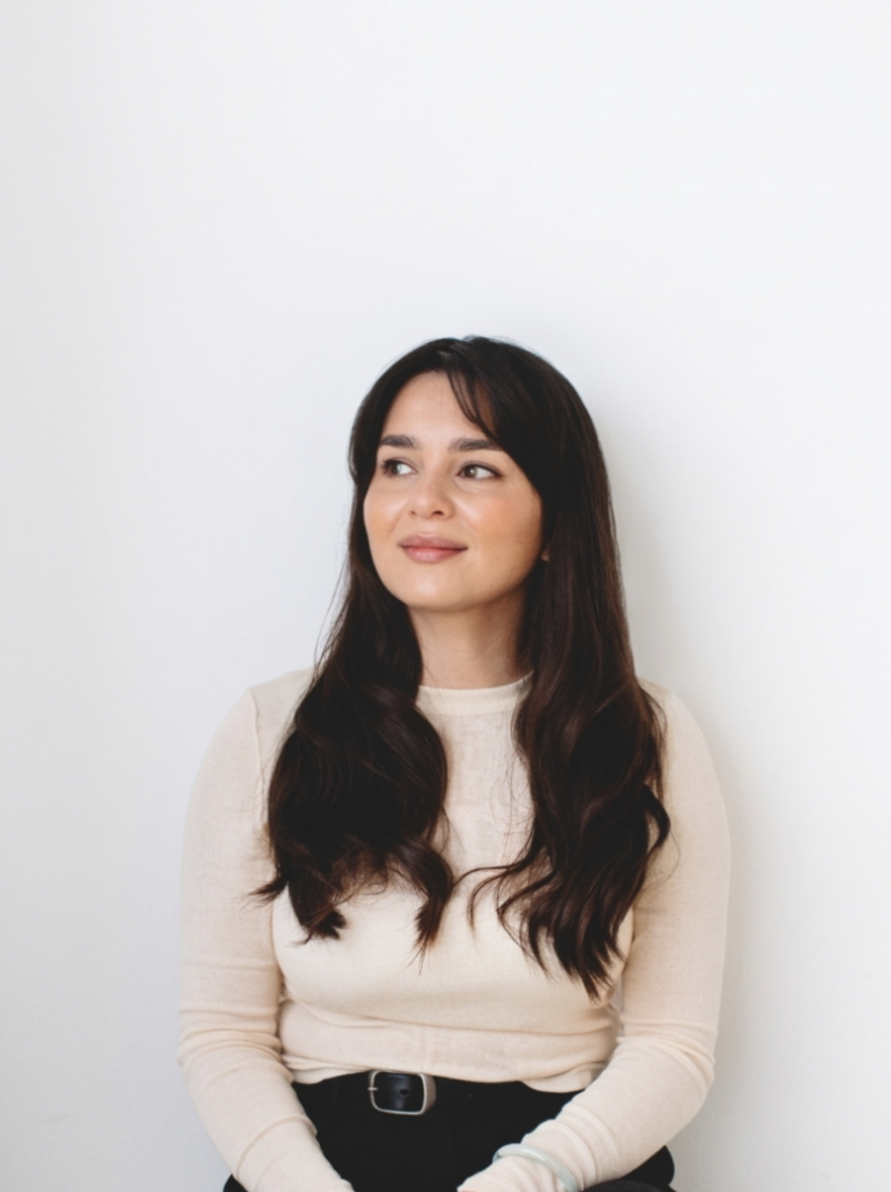 A young woman with long dark hair wearing a beige long-sleeve shirt and black pants, sitting against a plain white background.