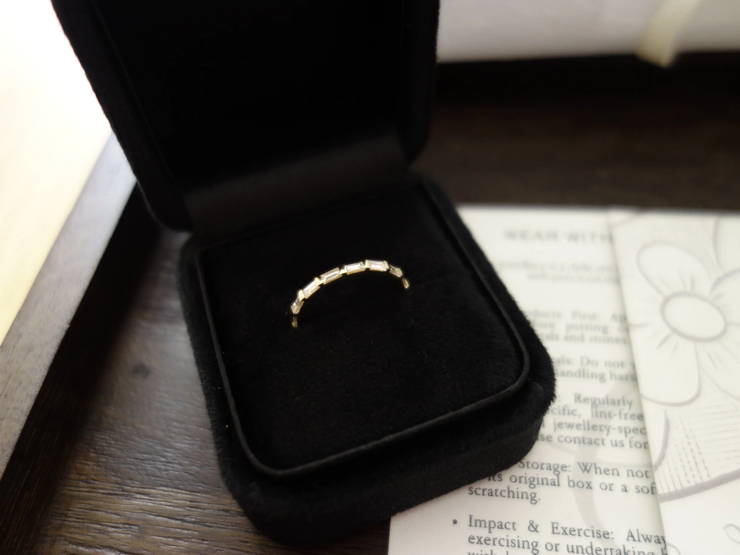 A gold bracelet with rectangular links in a black jewelry box resting on a wooden surface, with partially visible documents underneath.
