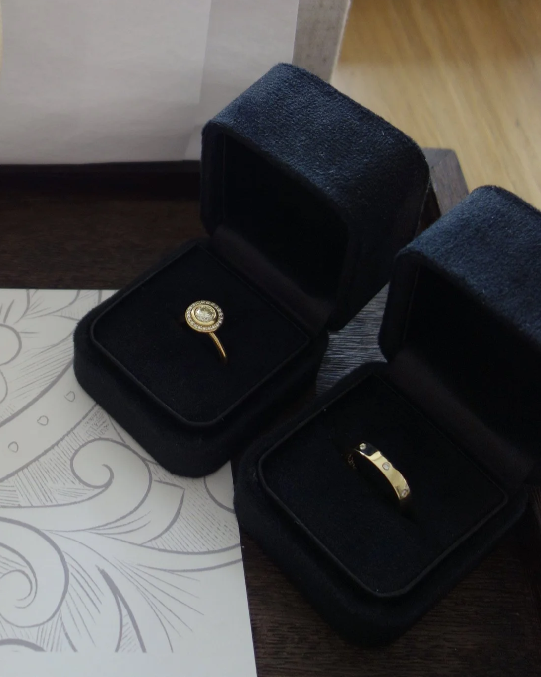 Two black jewelry boxes, each containing a ring, placed on a wooden surface with a decorative paper underneath.