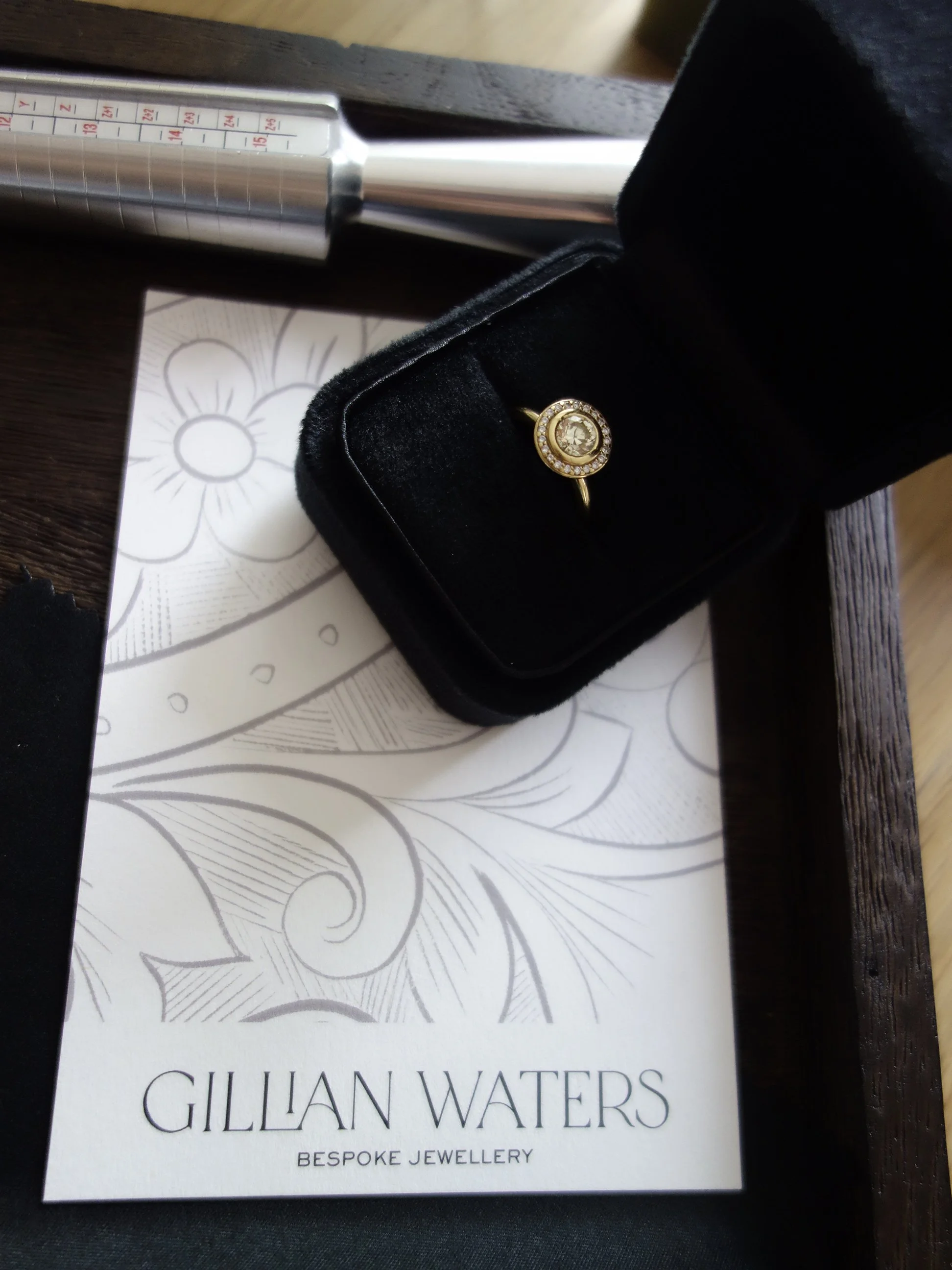 A gold ring with a large central diamond surrounded by smaller diamonds, placed inside a black jewelry box. The jewelry box is open, showing the ring. The box is on top of a white piece of paper or card with a floral design and the text 'GILLIAN WATERS BESPOKE JEWELLERY'.