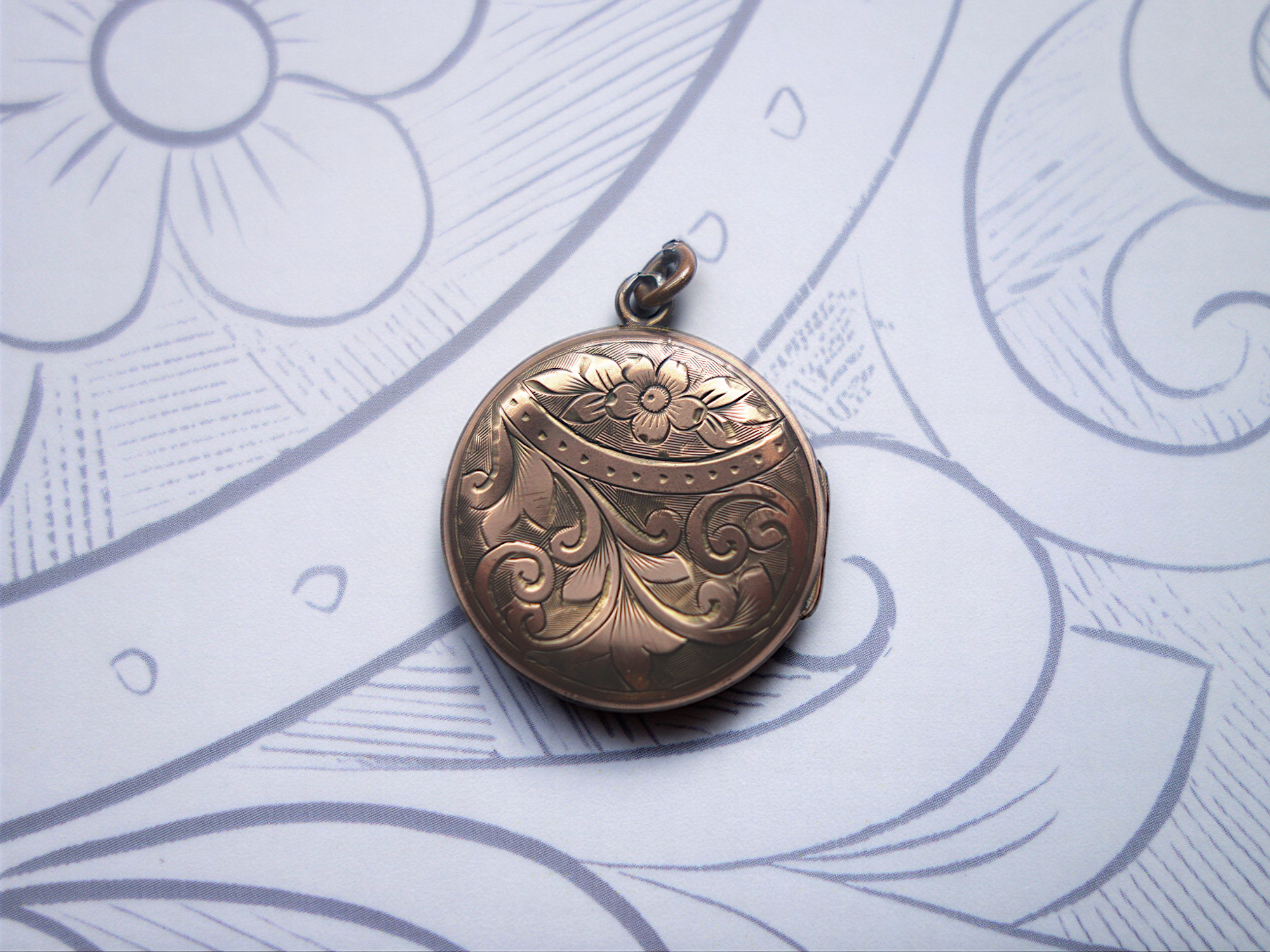A decorative gold pendant with floral and swirl engravings, laid on a sheet of paper with floral line art design.
