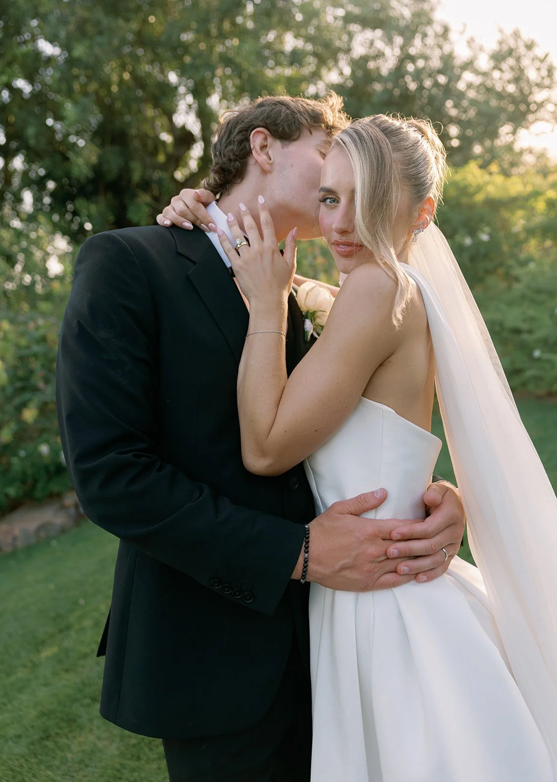 A bride and groom sharing a wedding kiss outdoors during sunset. The groom is in a black suit, and the bride wears a white strapless wedding dress and veil. They are embracing, with trees and greenery in the background.