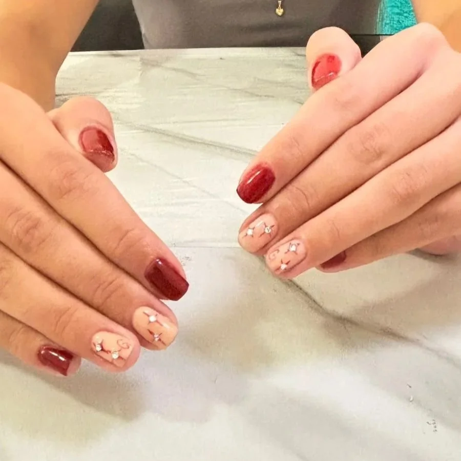 Manos con uñas pintadas de color vino y detalles de dibujos en las uñas con color beige y pequeños adornos blancos.