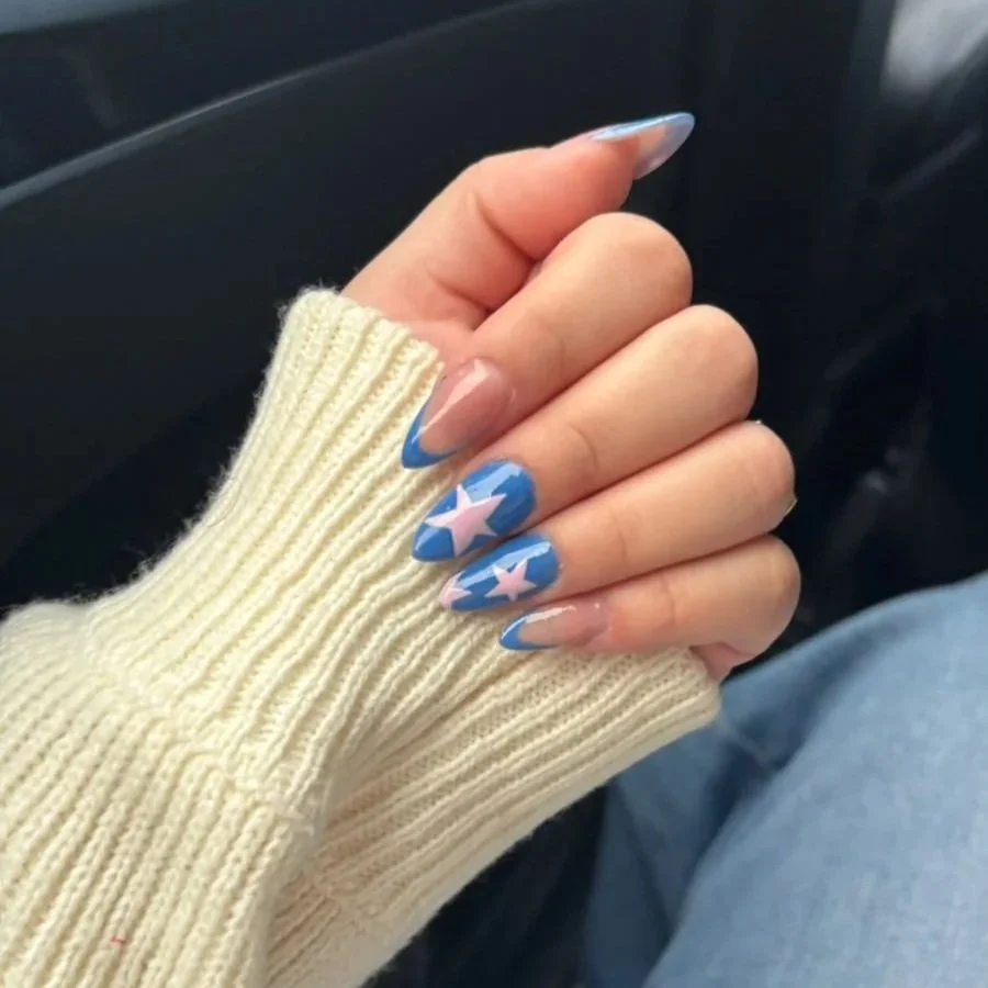 Uña con diseño de estrellas rosas y fondo azul, usando manicura en uñas largas, con un dedo envuelto en un guante de lana crema, en un vehículo.