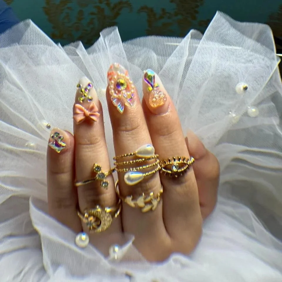 Mano con uñas decoradas con esmalte y adornos brillantes, usando varias joyas doradas y perlas en anillos, con una falda de tul blanca y cinta con perlas.