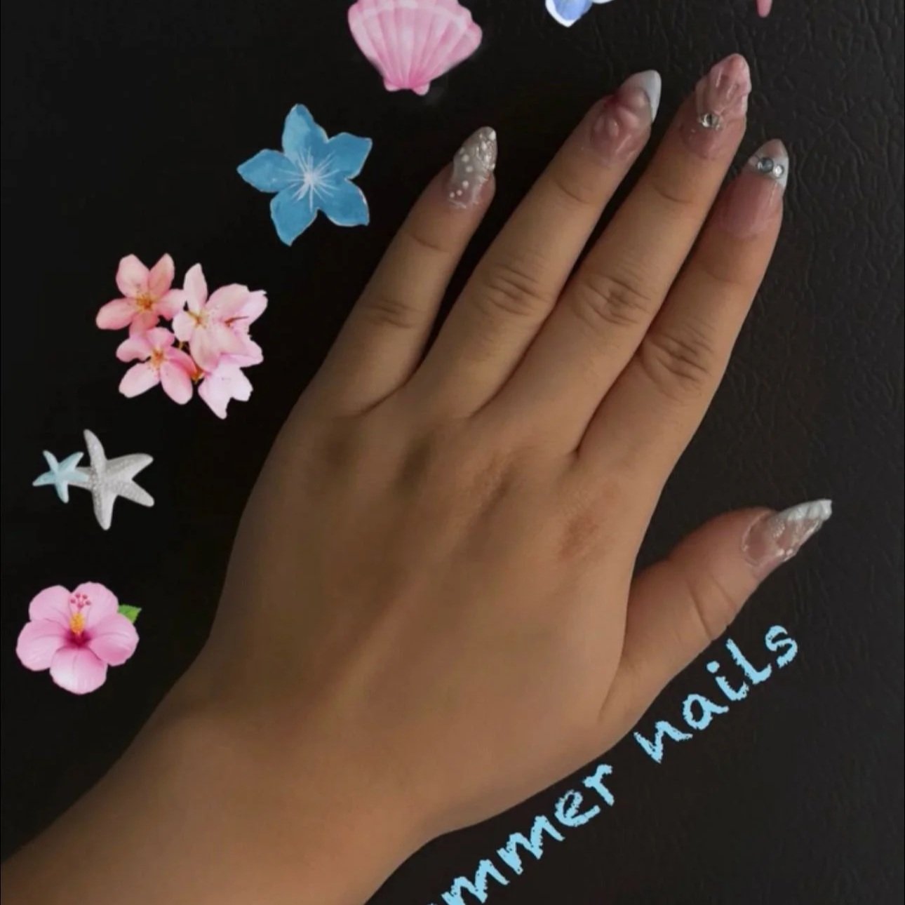 Mano con uñas decoradas con diseño en tonos blancos y accesorios brillantes, sobre fondo negro con flores de colores claros y la frase 'summer nails' en la esquina inferior derecha.