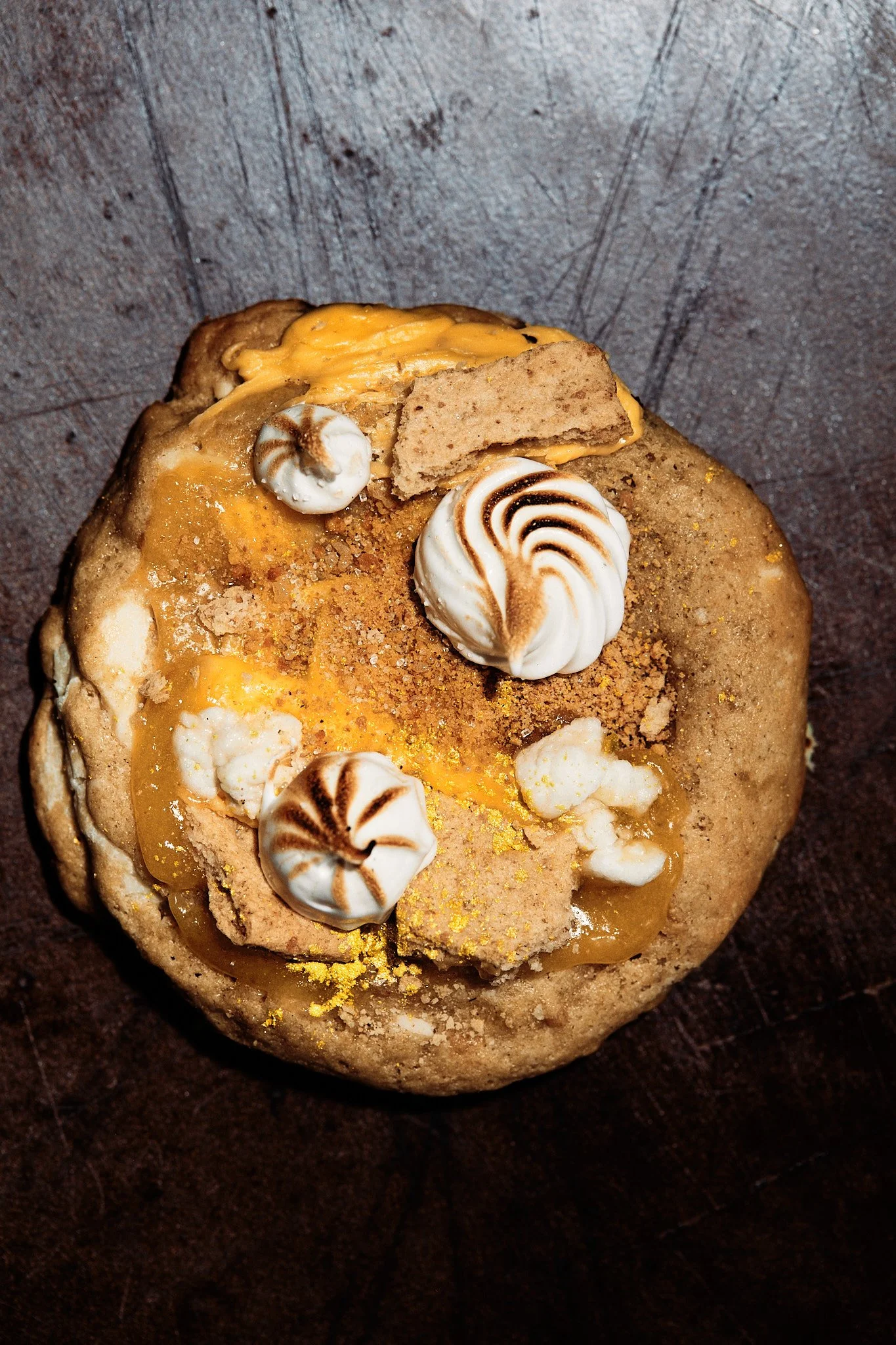 A dessert topped with toasted meringue, crushed cookies, lemon curd, and whipped cream garnished with lemon zest on a dark wooden surface.