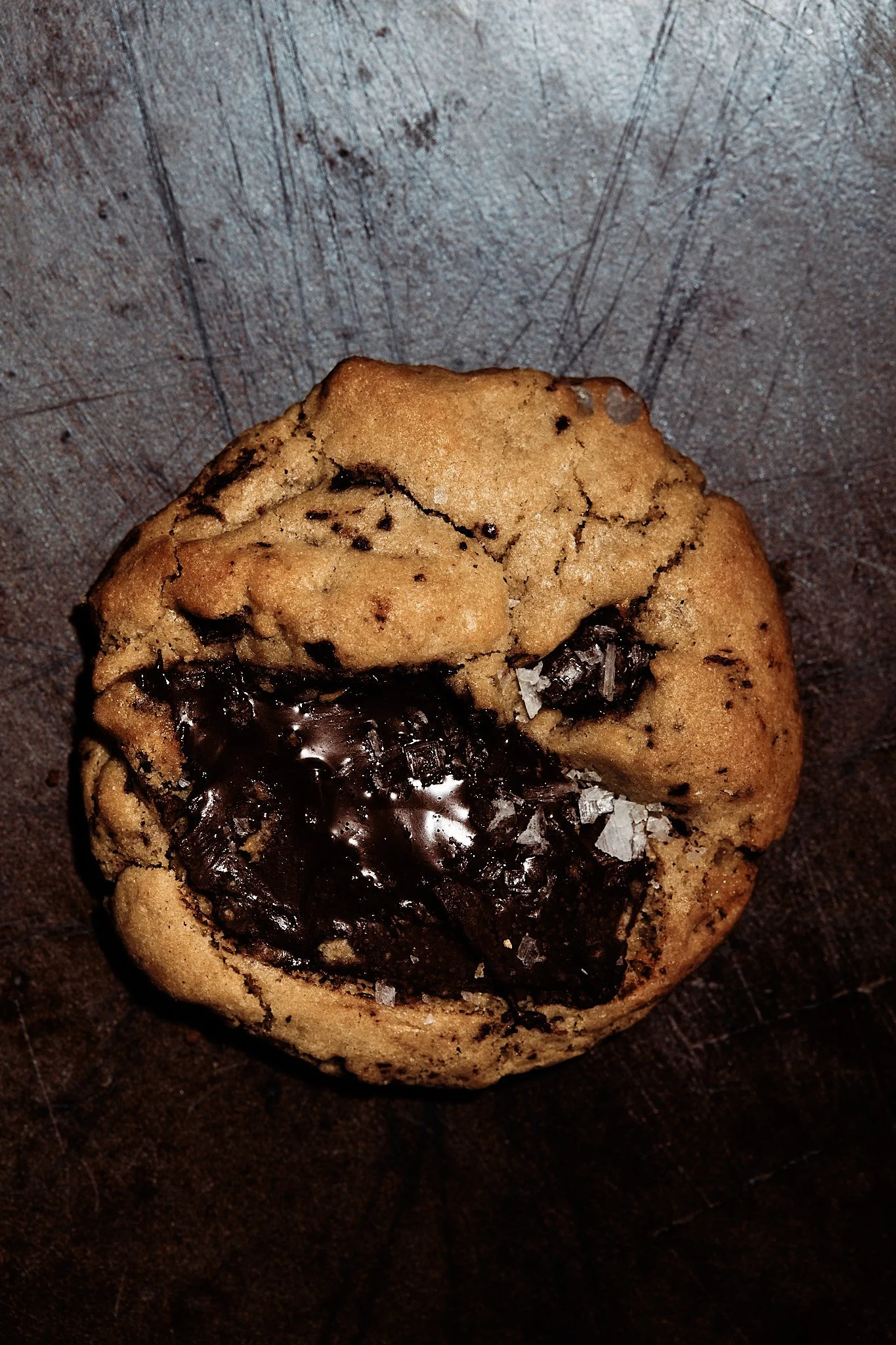Close-up of a chocolate chip cookie with melted chocolate and salt flakes on a dark surface.