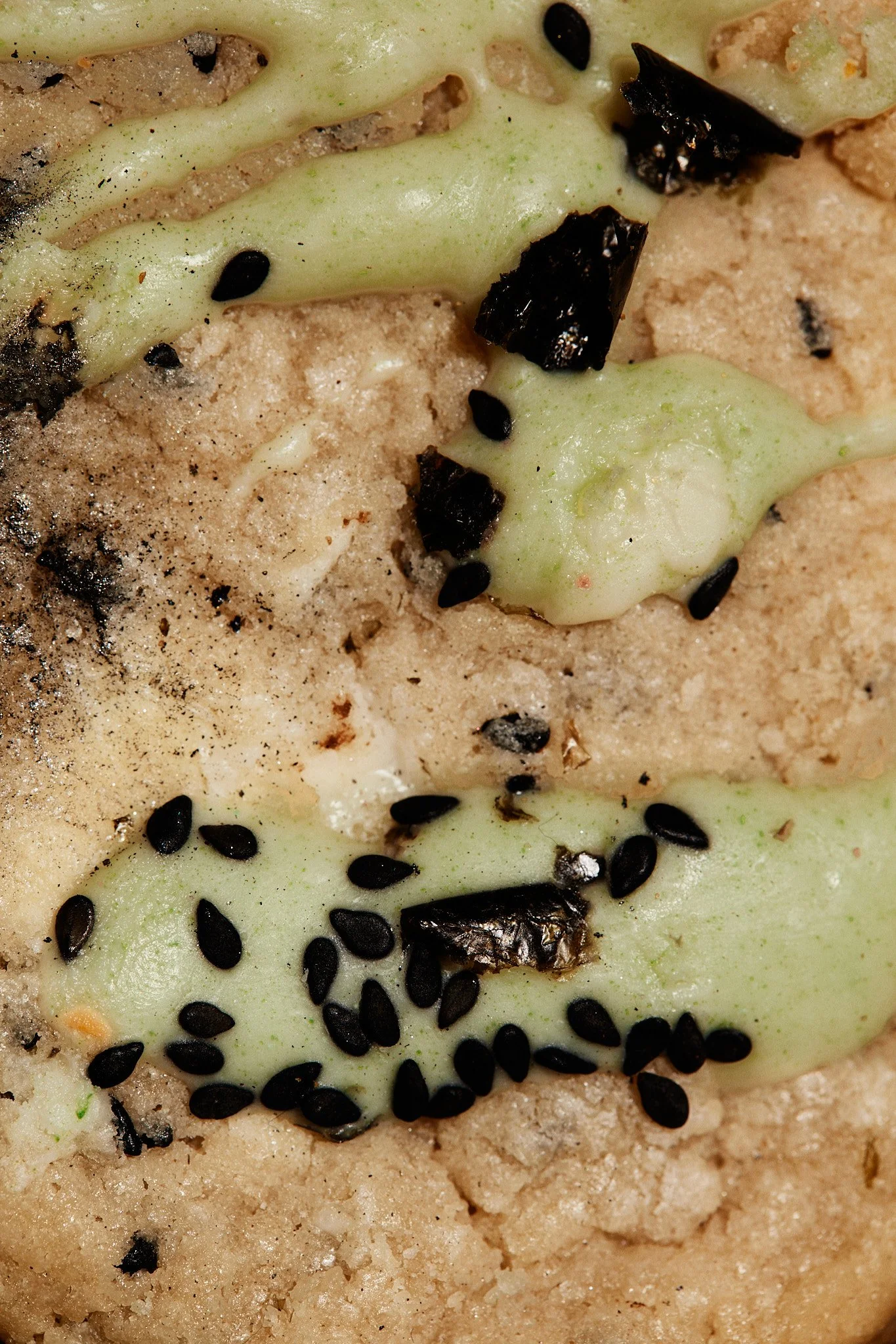 Close-up of a baked dessert with a crust, topped with greenish cream, black sesame seeds, and small chocolate pieces.