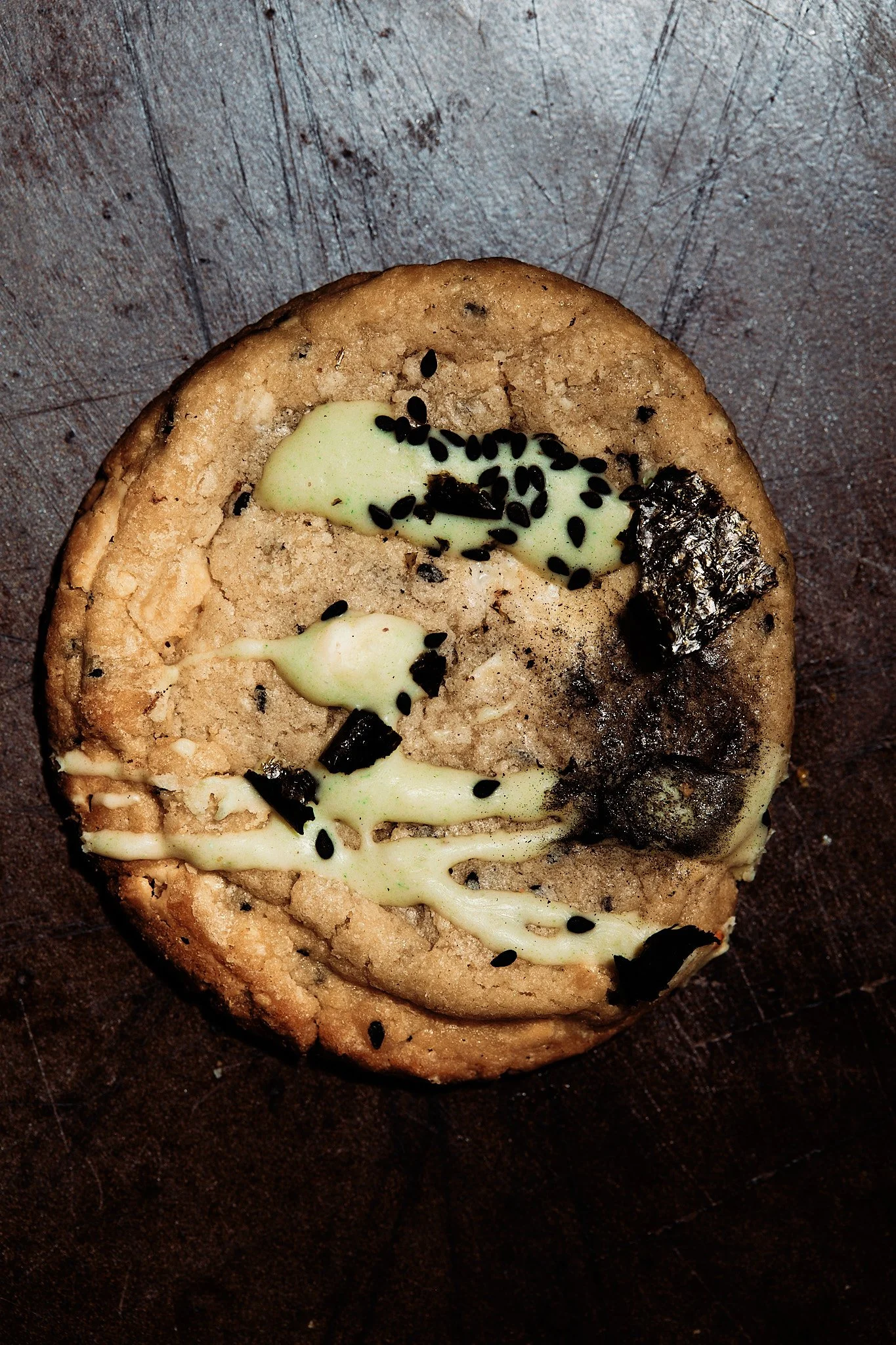 Close-up of a broken cookie with vanilla glaze, black sesame seeds, and edible silver on a dark wooden surface.