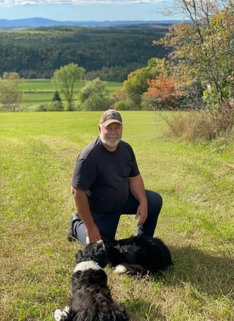 A man kneeling on one knee in a grassy field with two black and white puppies, natural landscape with trees and hills in the background.