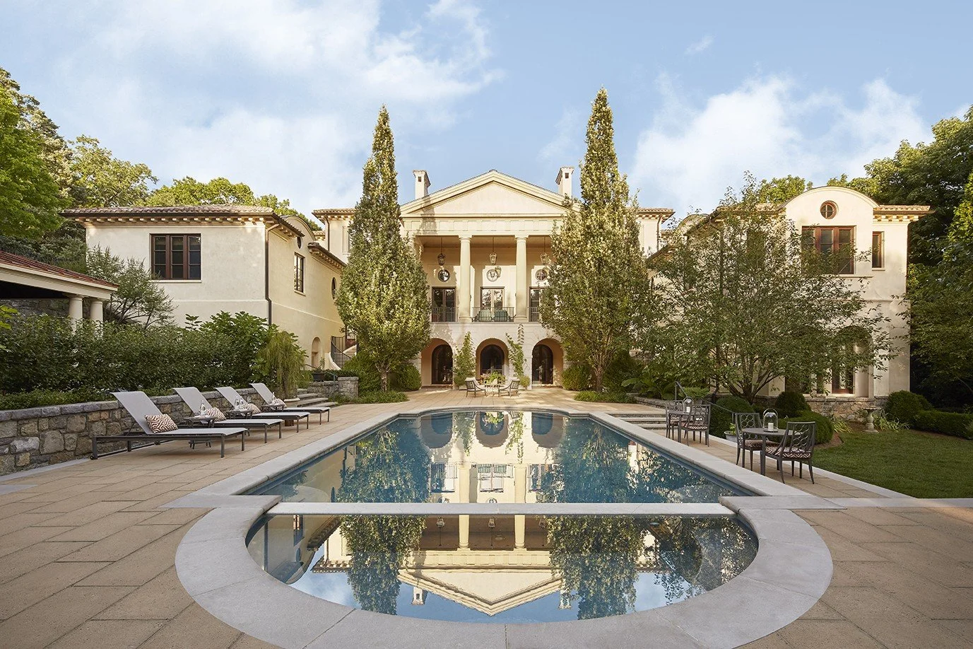 A large white mansion with a symmetrical, classical design, situated behind a rectangular swimming pool with a circular hot tub, surrounded by chairs, tables, and lush green trees under a blue sky with some clouds.