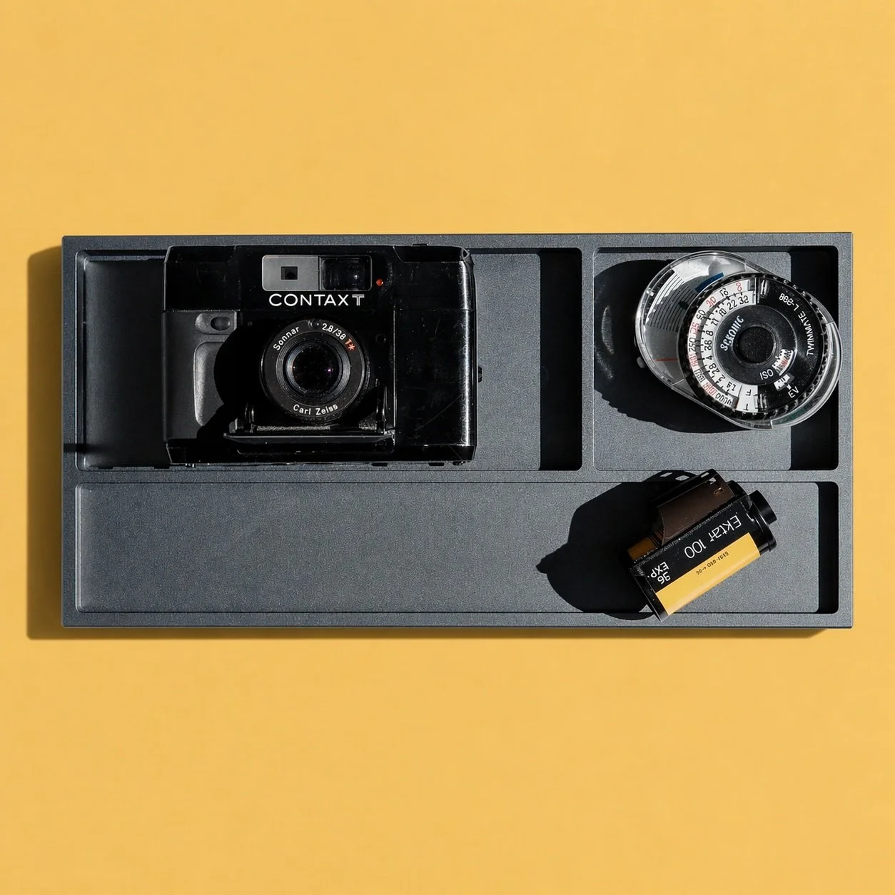 A vintage Contax T camera, a light meter, and a roll of Kodak Ektar 100 film on a yellow background.