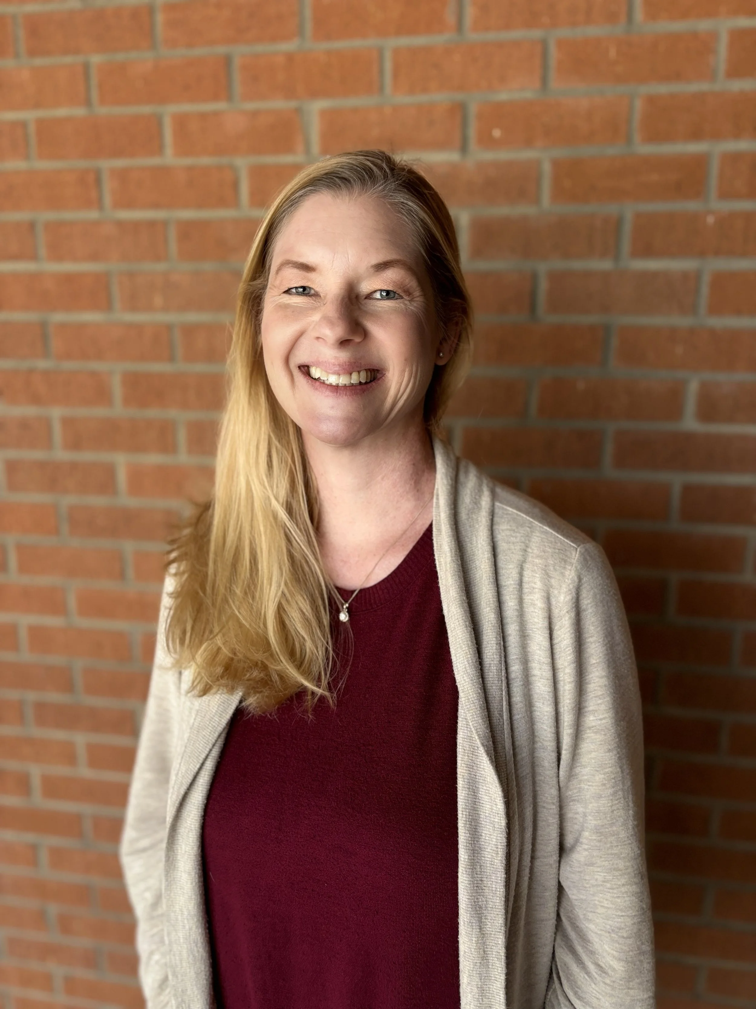 A smiling woman with long blonde hair standing in front of a red brick wall, wearing a beige cardigan over a maroon top.