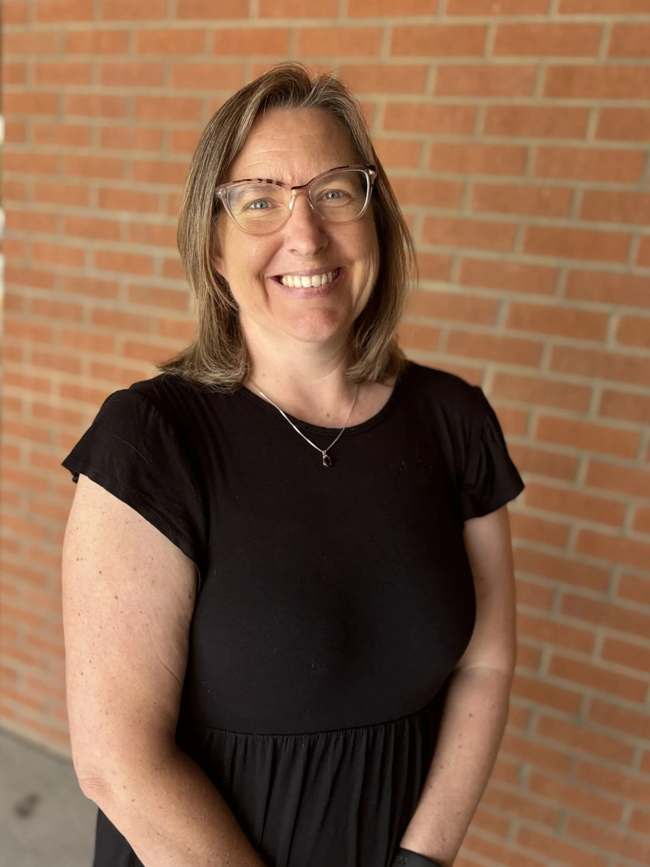 Close-up of a woman with shoulder-length brown hair and glasses, smiling, wearing a black dress with ruffled sleeves and a silver necklace, standing against a red brick wall.