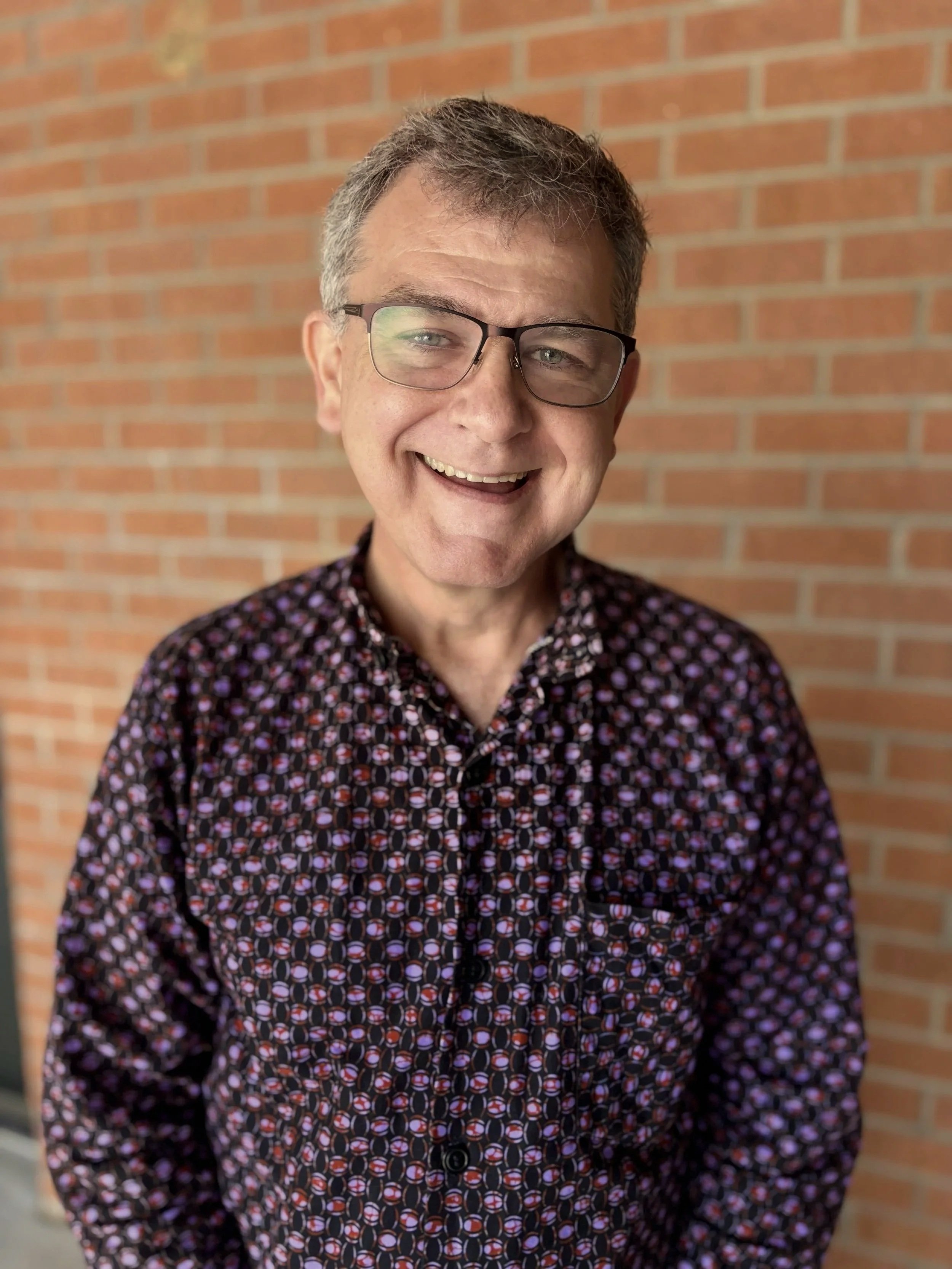 A man with gray hair, glasses, and a patterned shirt smiling in front of a brick wall.