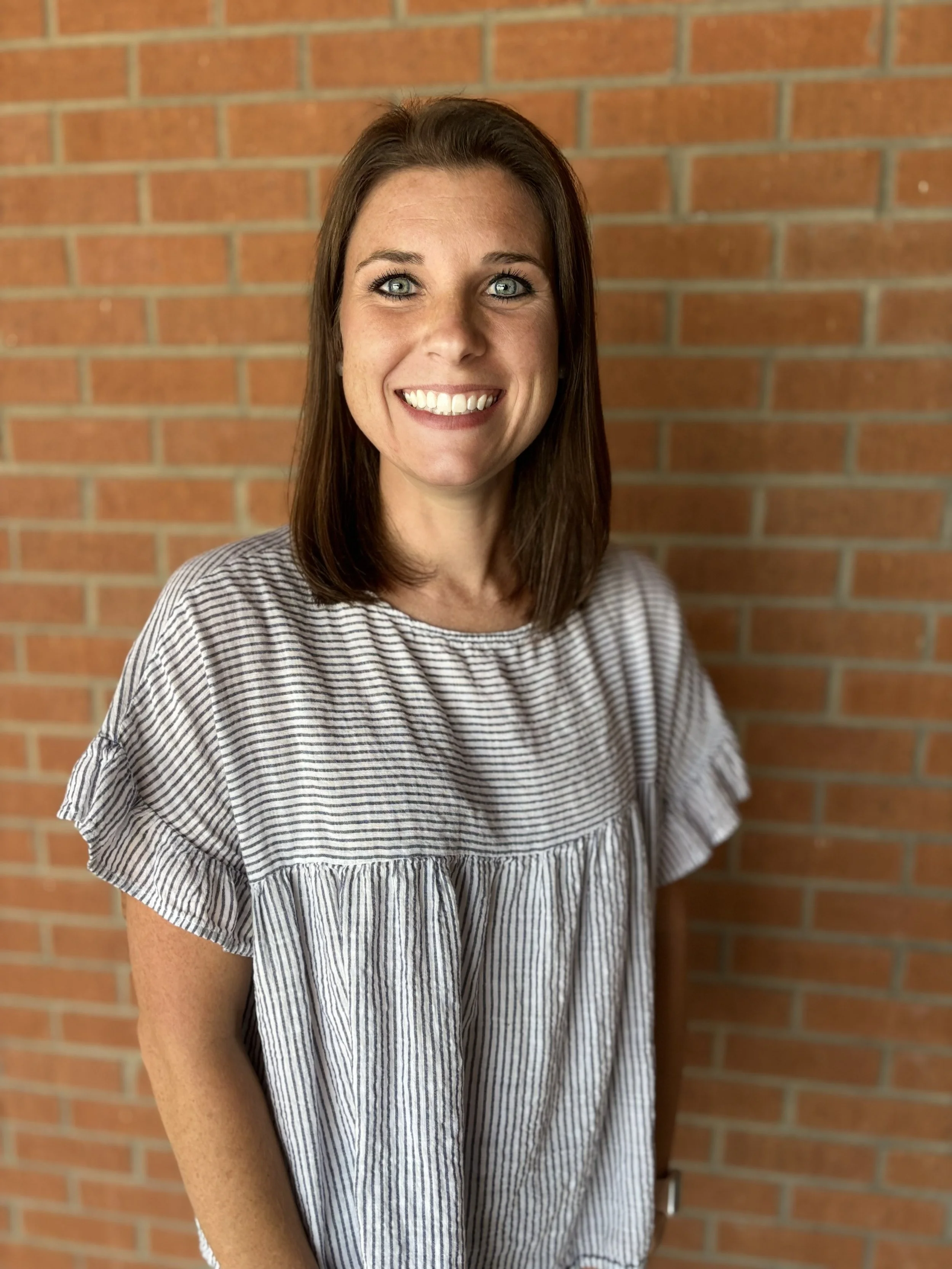 A woman with medium-length brown hair and blue eyes smiling at the camera, wearing a striped gray and white ruffled top, standing in front of a brick wall.