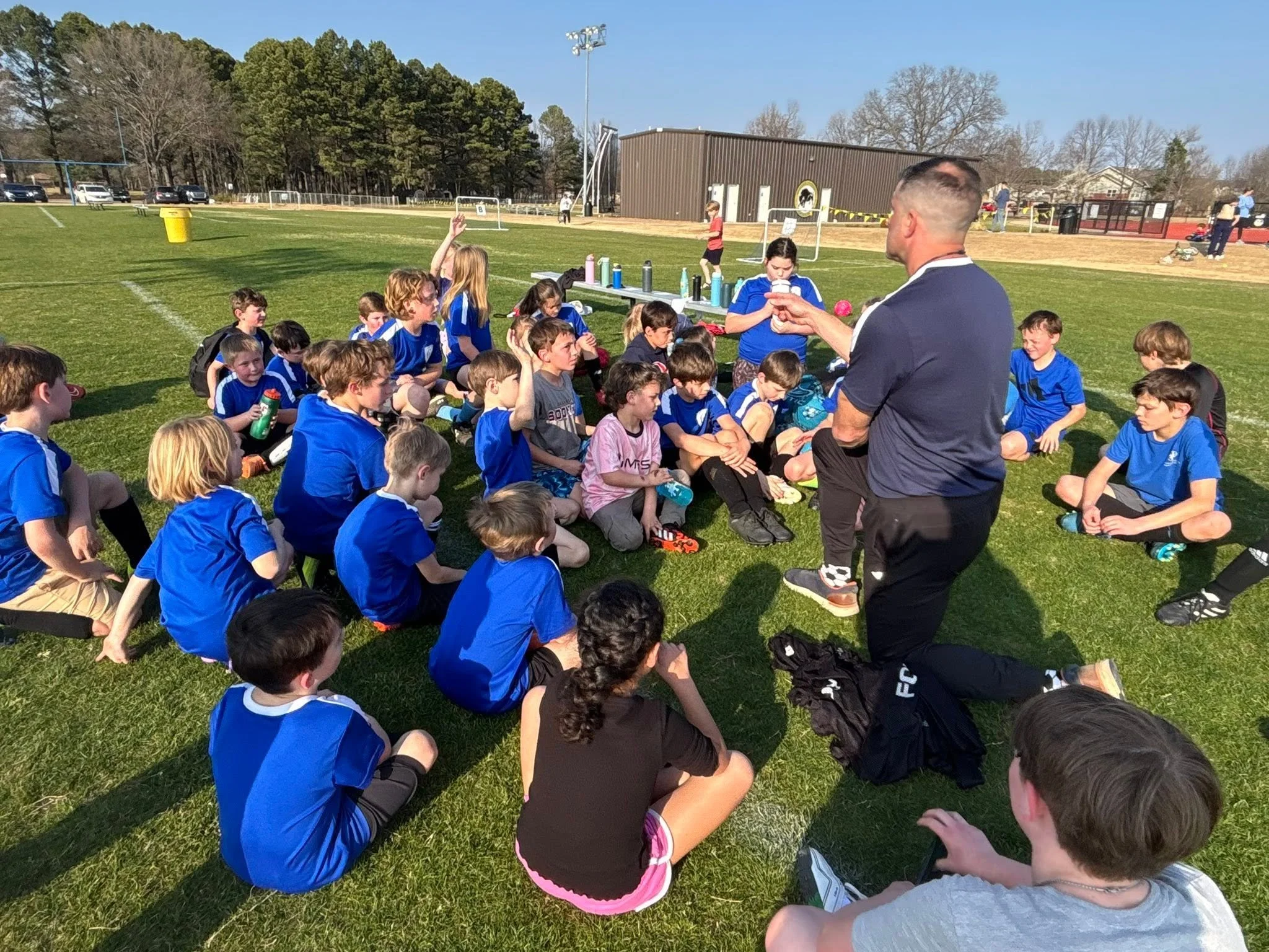 A group of young children dressed in blue sports jerseys sitting on the grass for a break during a soccer game or practice, with a coach talking to them, in a park or sports field on a sunny day.