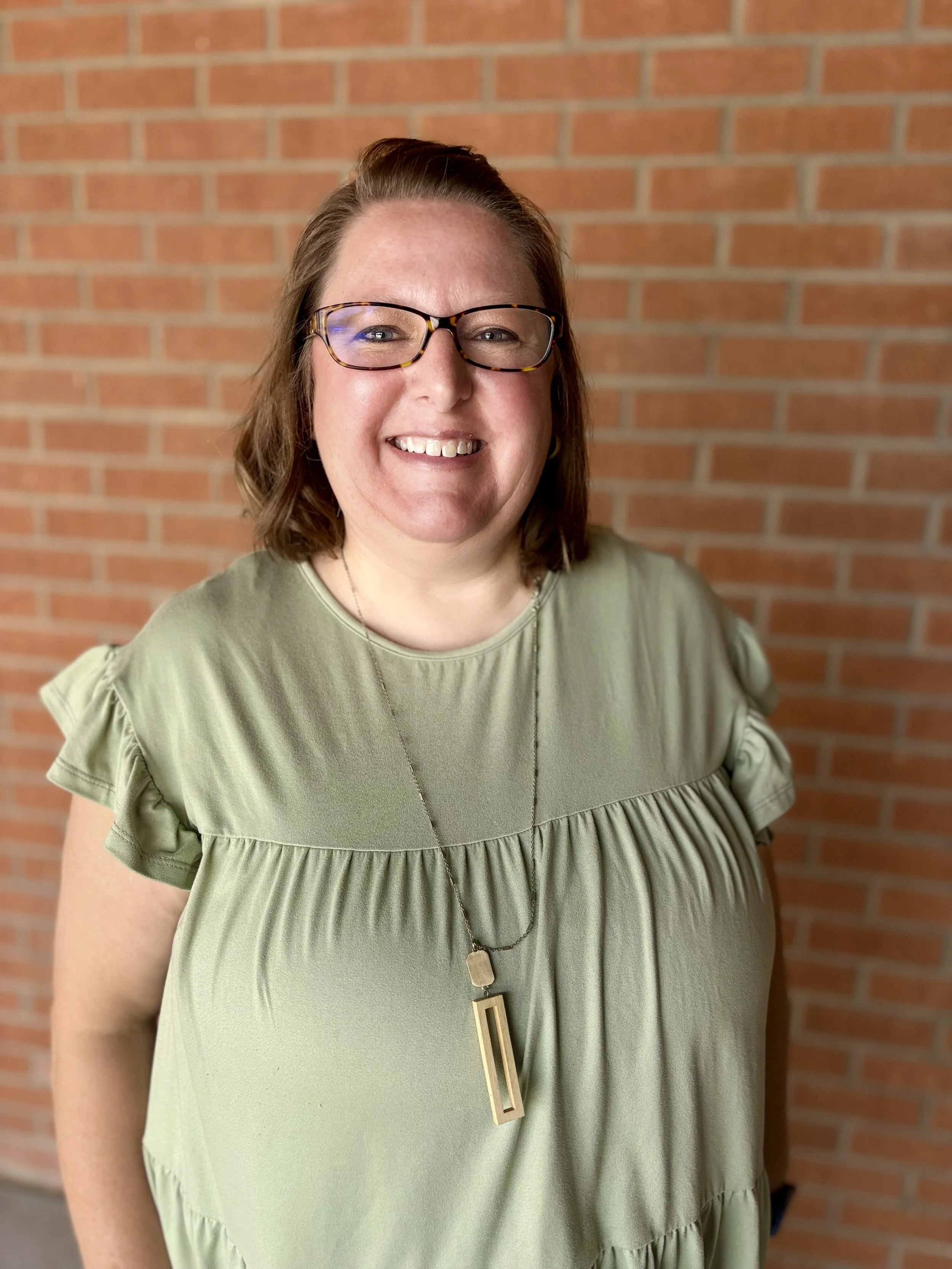 Close-up of a woman with glasses smiling, wearing a green dress with ruffled sleeves and a long pendant necklace, standing in front of a brick wall.